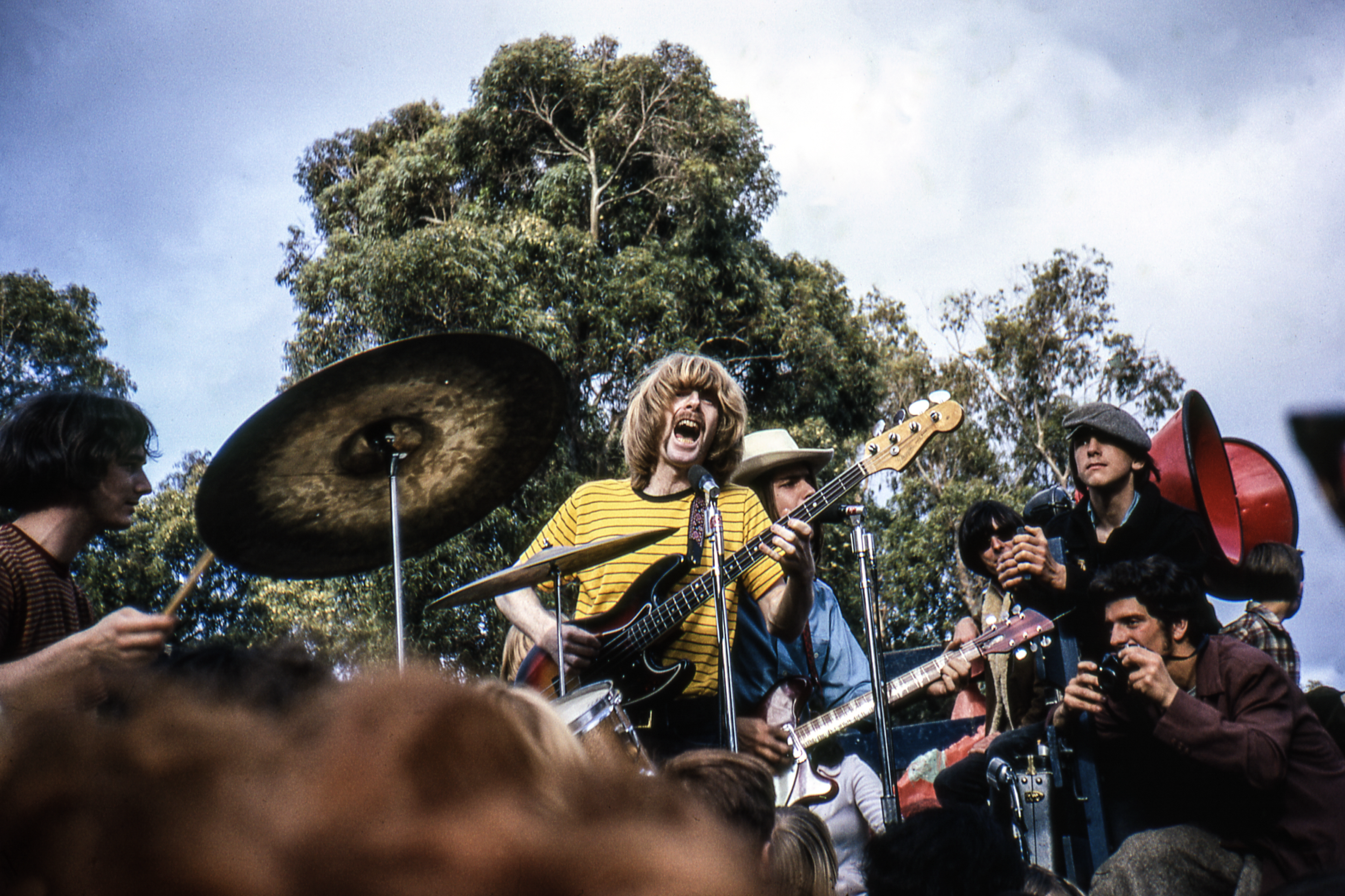 A band performs energetically outdoors, with a drummer, guitarist, and bassist in focus. Trees and a cloudy sky are in the background, and a crowd watches closely.