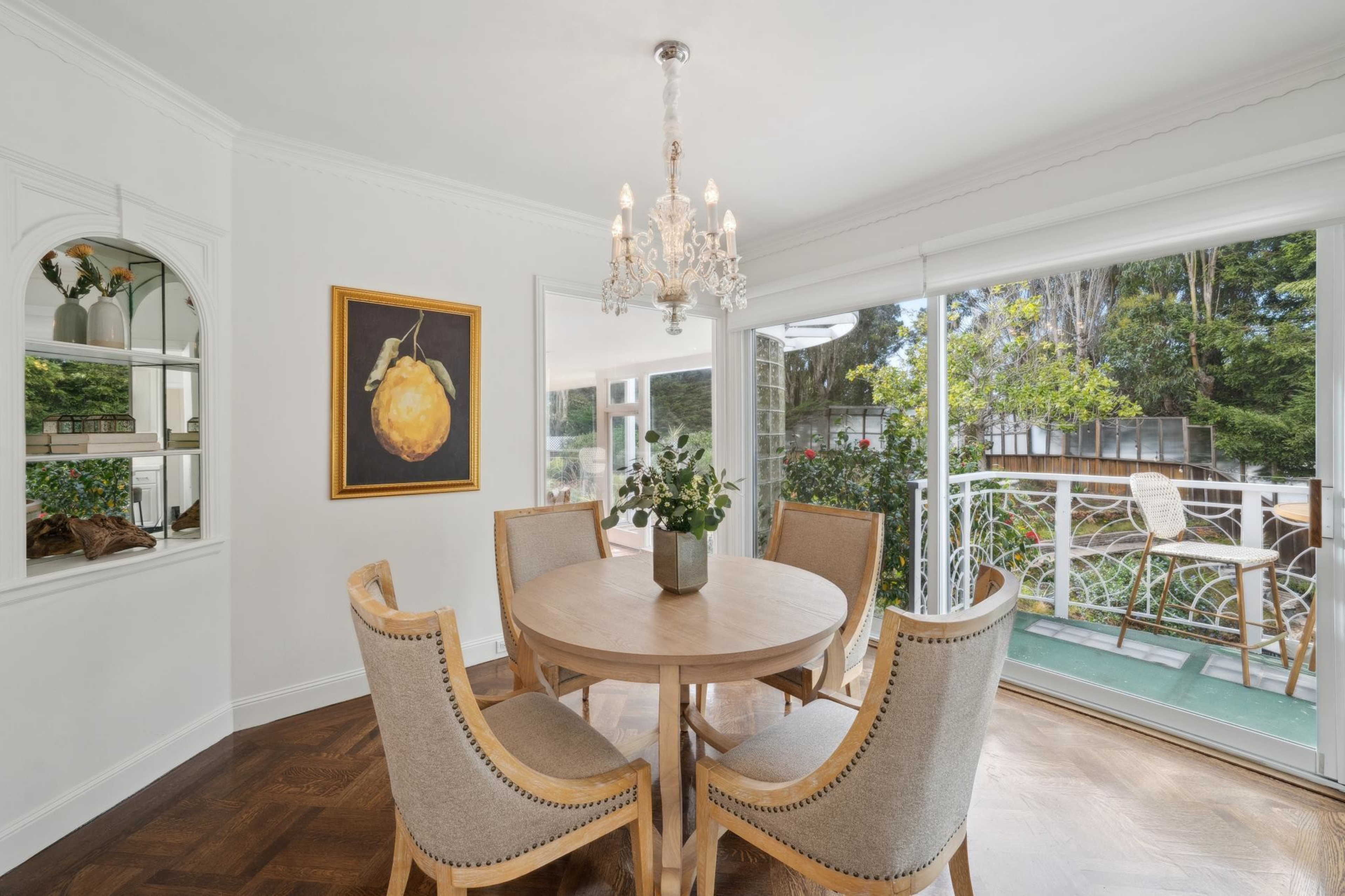 A bright dining area features a round wooden table with five beige cushioned chairs, a chandelier, a fruit painting, and large sliding glass doors opening to a balcony.