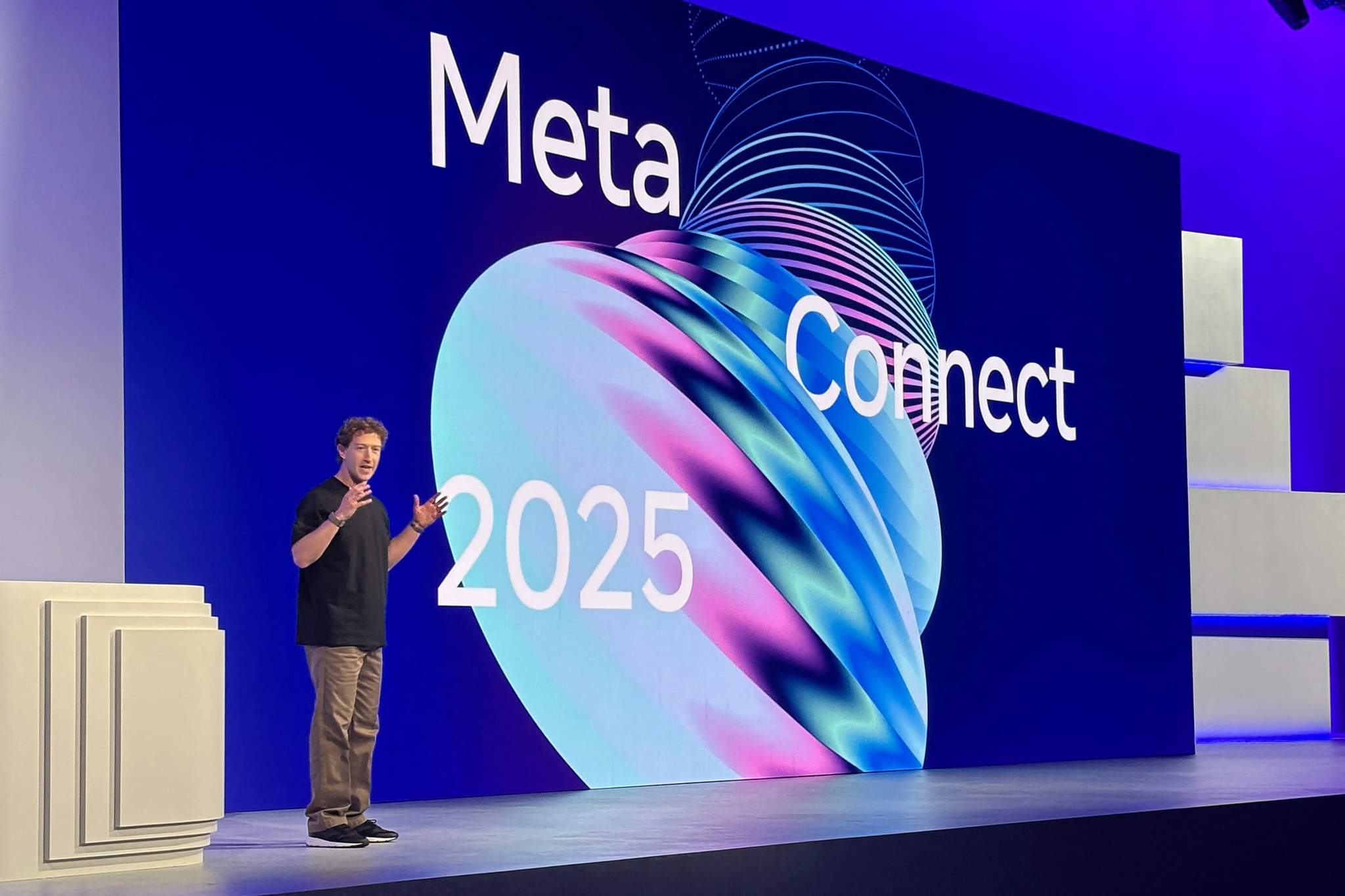 A man stands on stage giving a presentation in front of a large screen displaying "Meta Connect 2025" with colorful abstract shapes on a dark blue background.