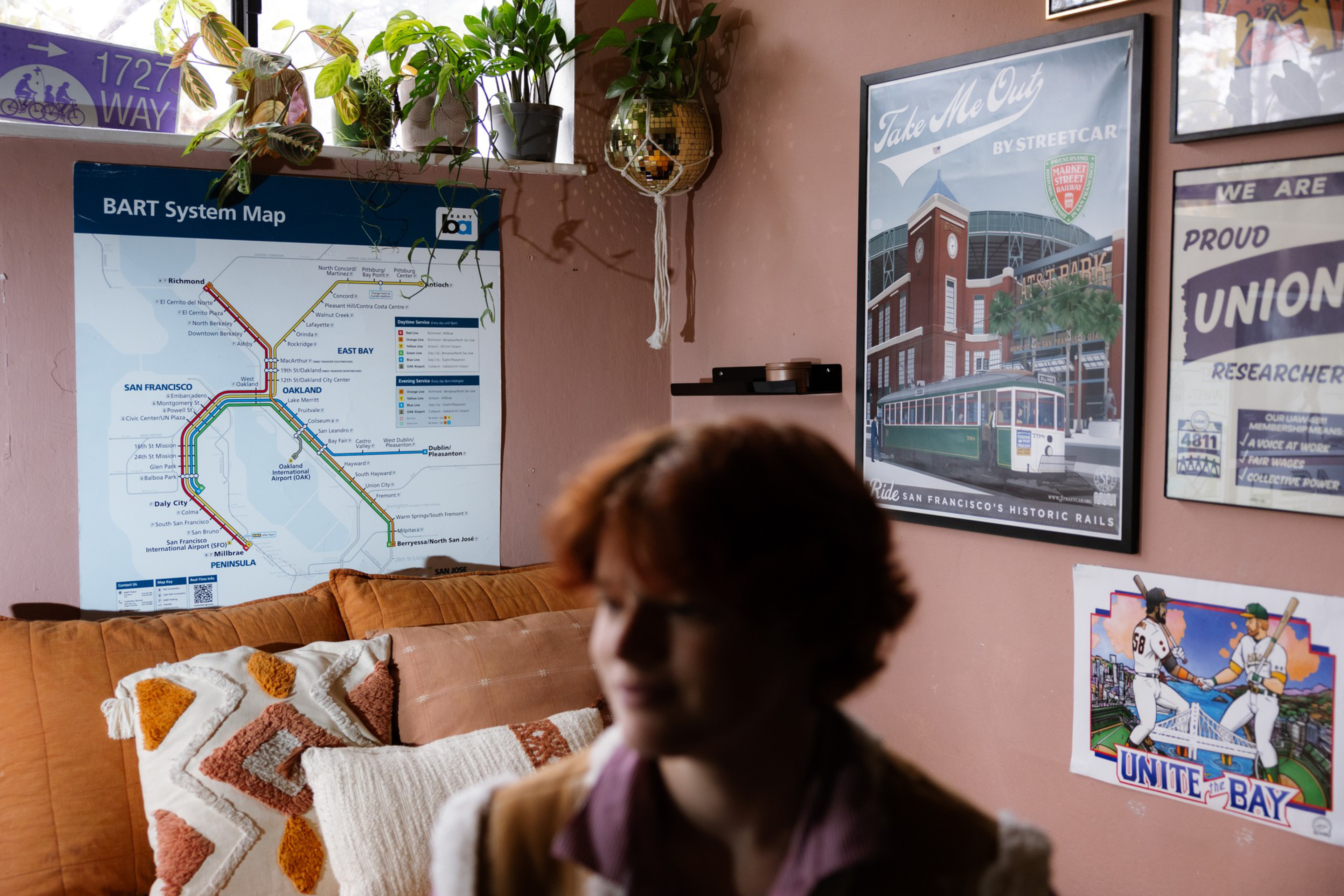 A cozy room corner with a tan couch, patterned pillows, hanging plants, and framed posters including a BART map and vintage streetcar ad.