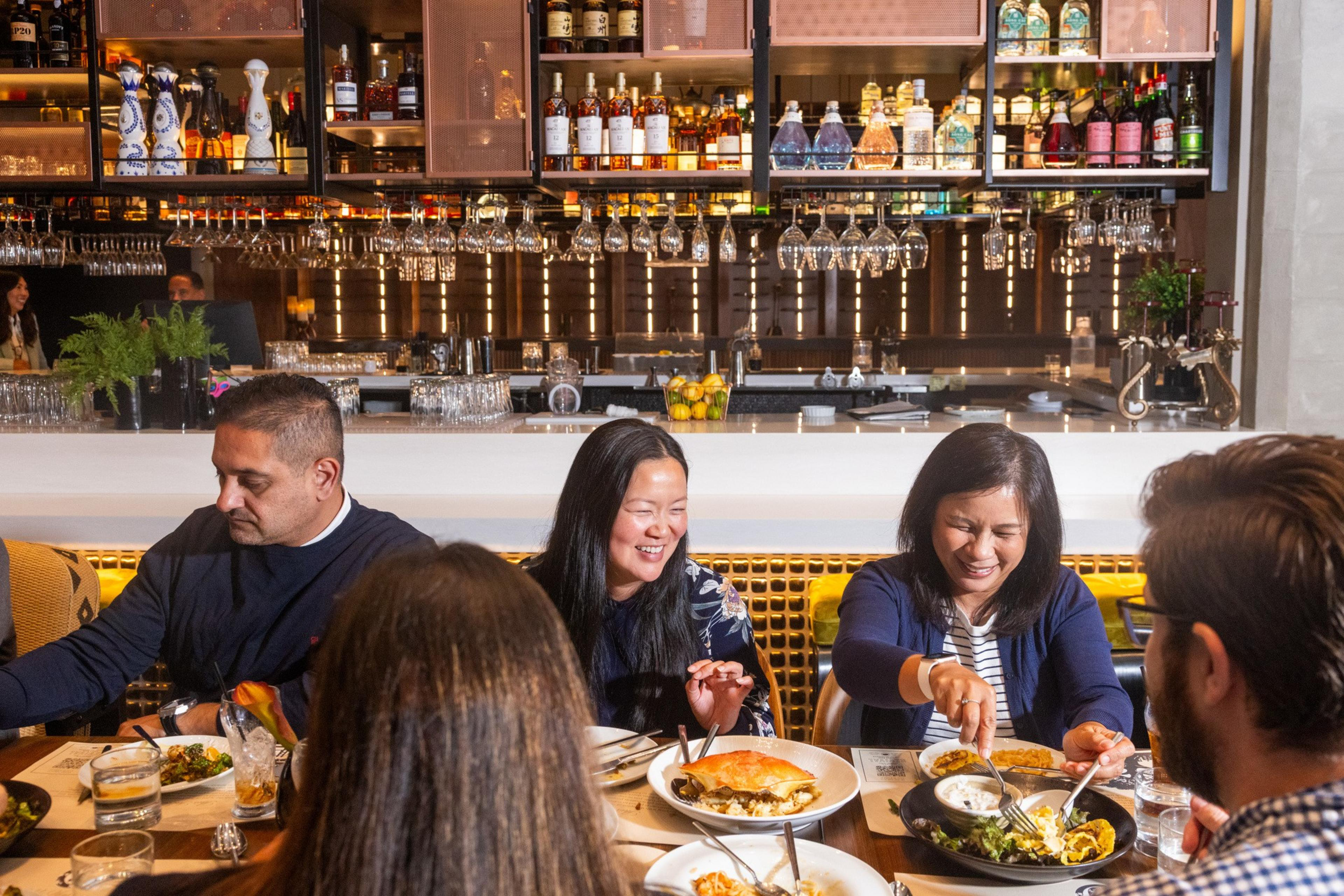 Five people sit around a table in a lively restaurant, enjoying diverse dishes and drinks with smiles and engaged conversation.