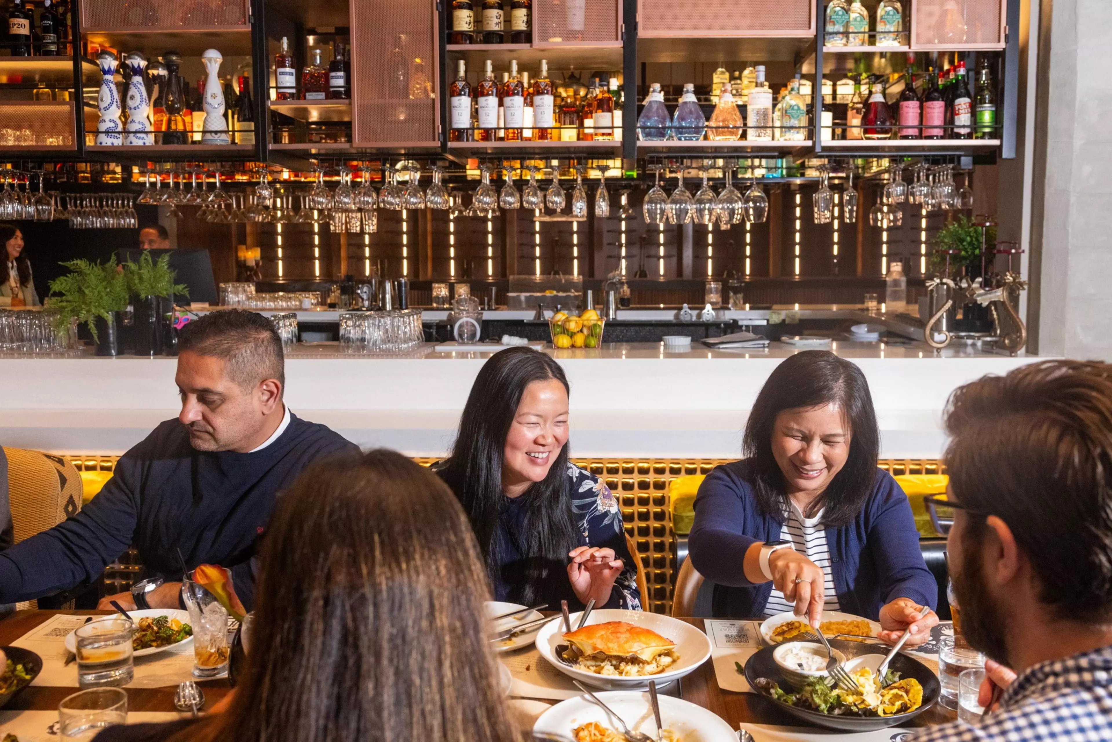 Five people sit around a table in a lively restaurant, enjoying diverse dishes and drinks with smiles and engaged conversation.