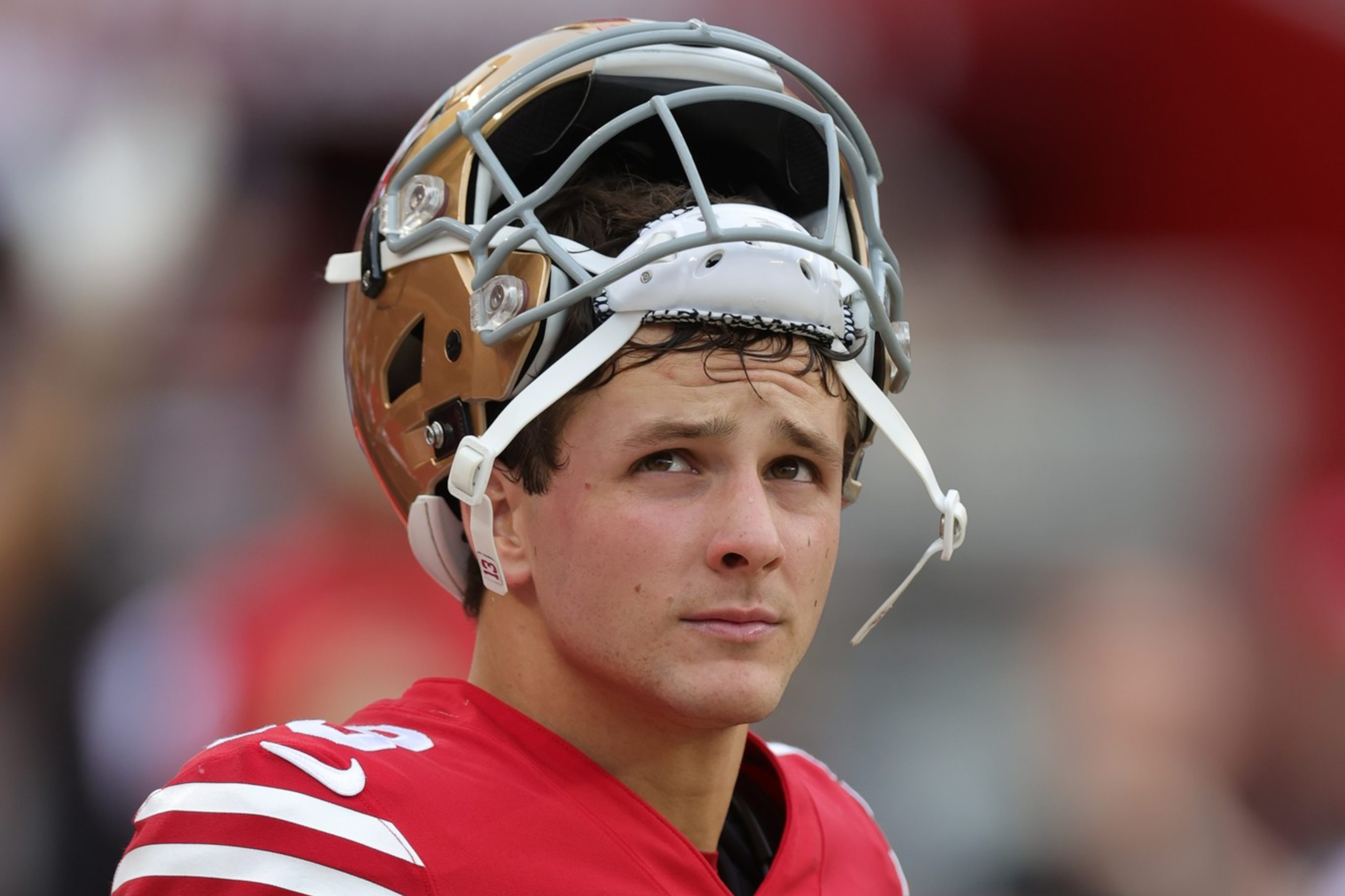 A football player wearing a red and white uniform stands with his helmet resting on top of his head, looking slightly upward and to the side.