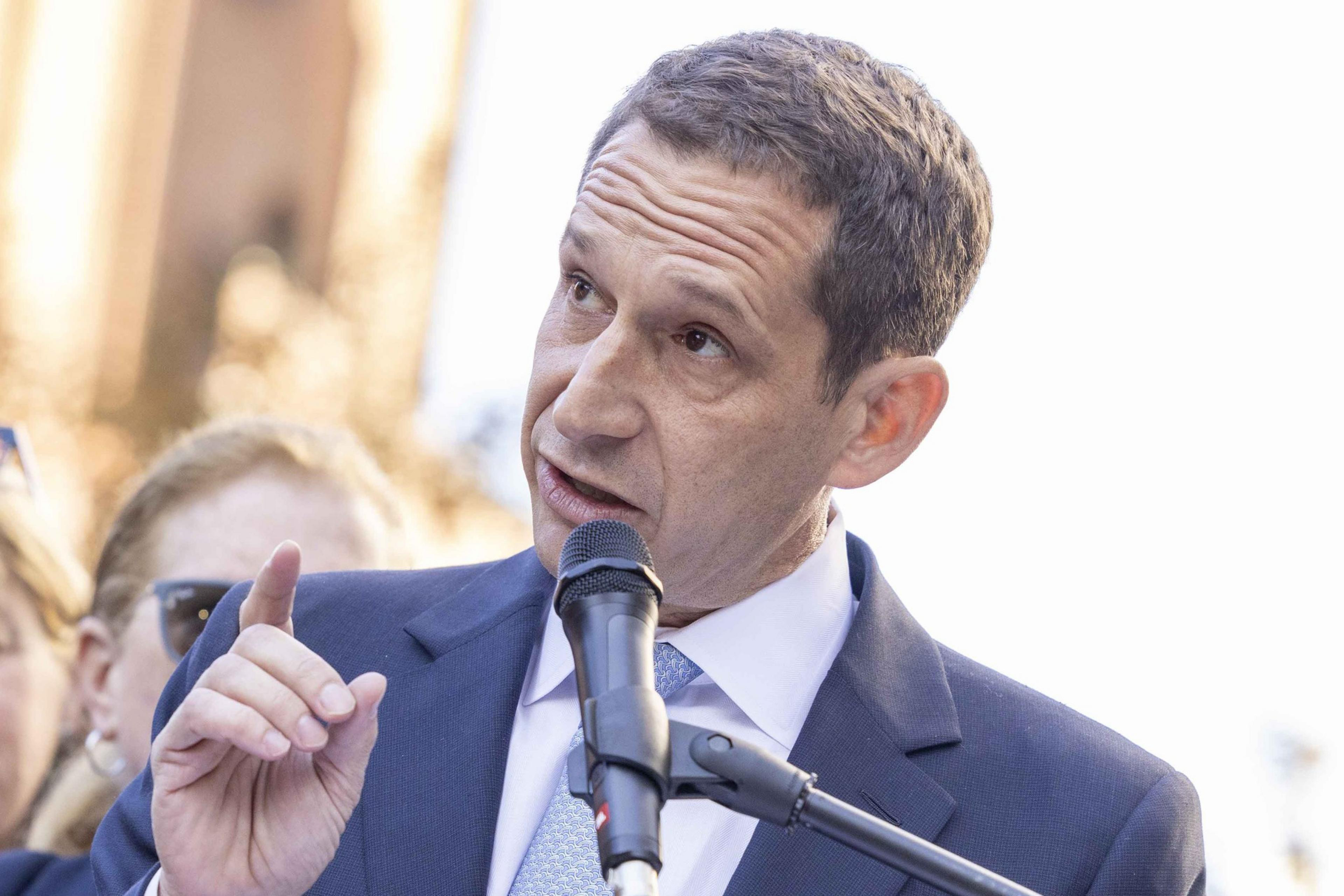 A man in a suit speaks into a microphone, gesturing with one hand. The background is slightly blurred, suggesting an outdoor setting with people.