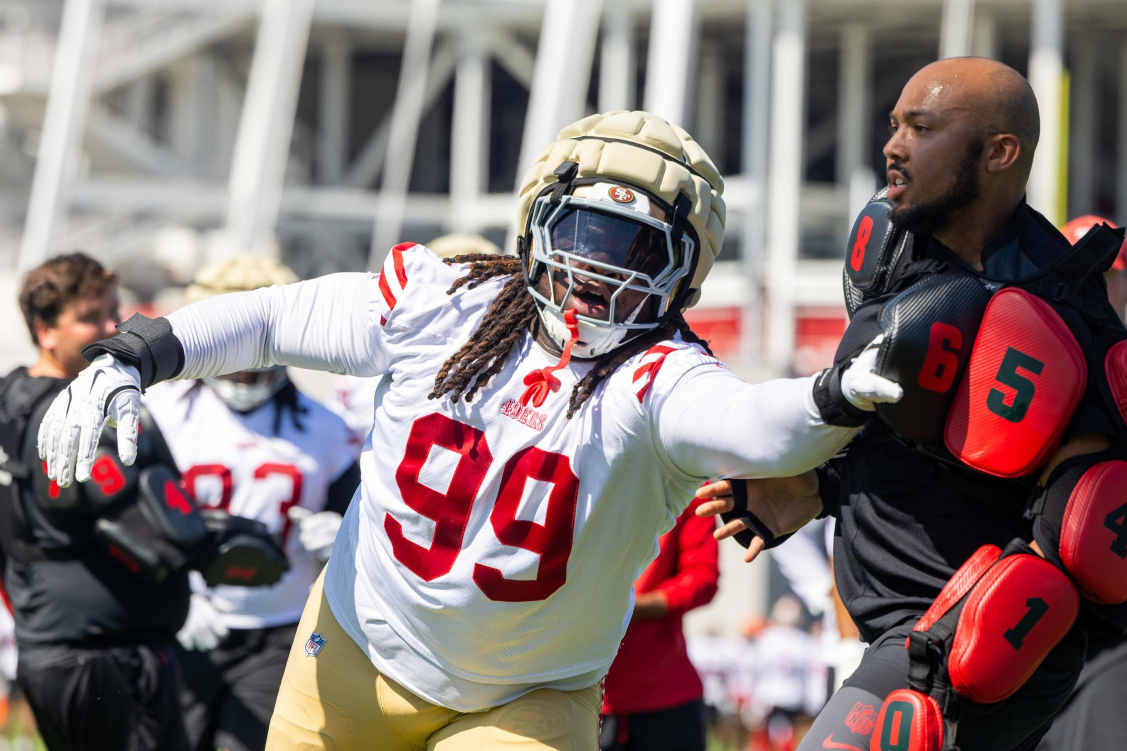 A football player in a white and red uniform with number 99 practices by pushing against a padded opponent. Other players and a building are in the background.