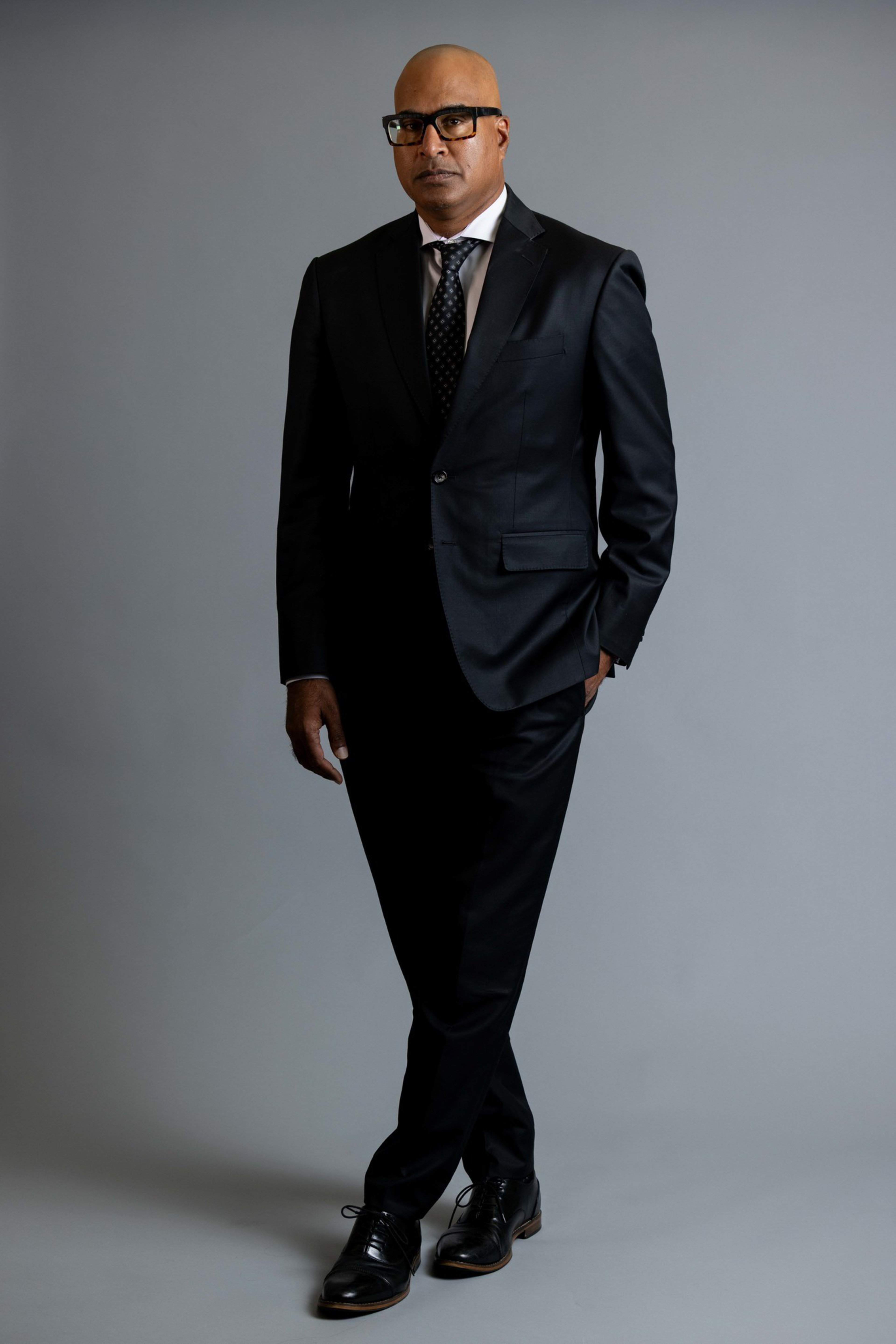 A bald man wearing black glasses, a black suit, white shirt, and black tie stands with one hand in his pocket and legs crossed against a gray background.