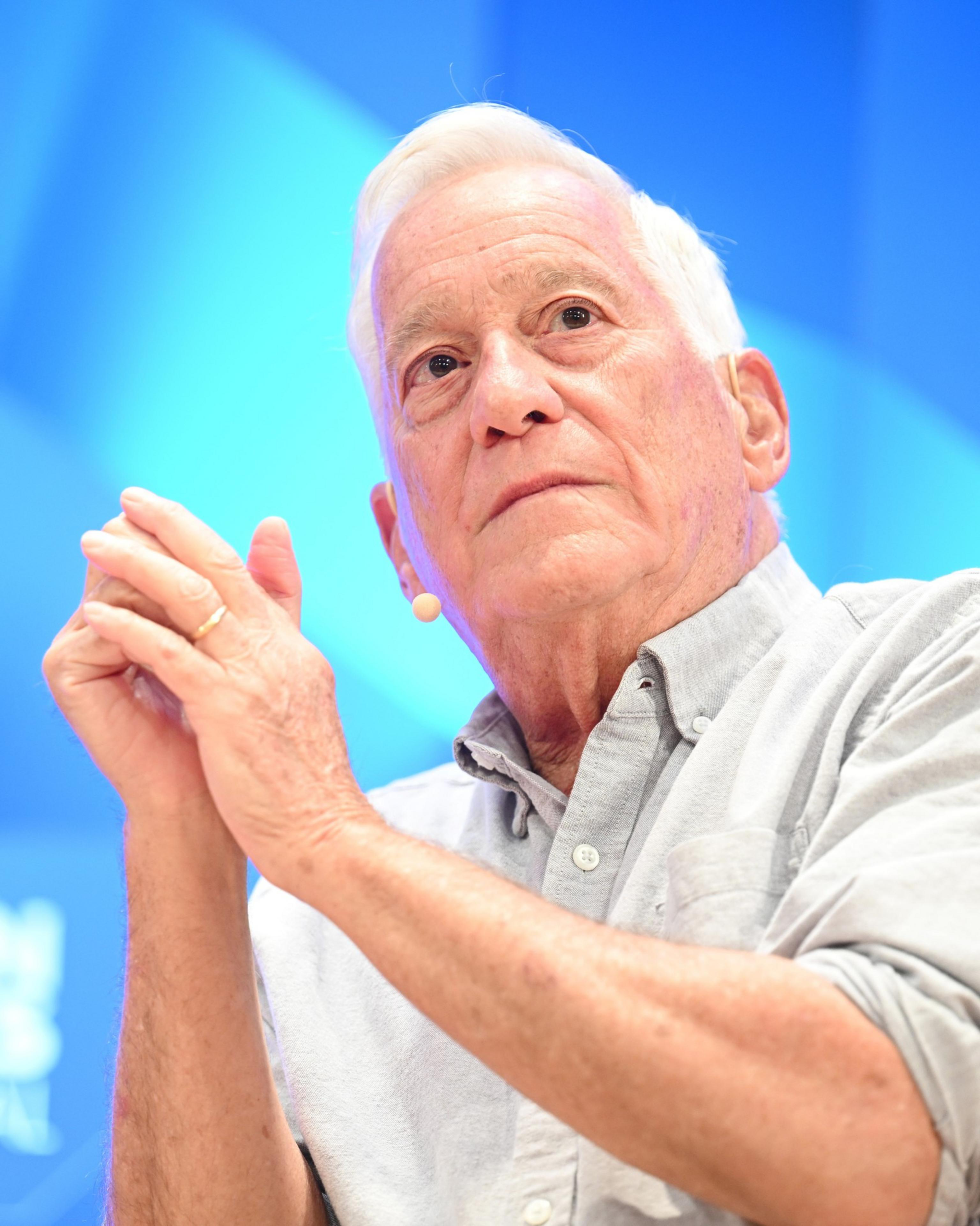 An elderly man with white hair and a light gray button-up shirt clasps his hands, looking thoughtfully to the side against a blue background.