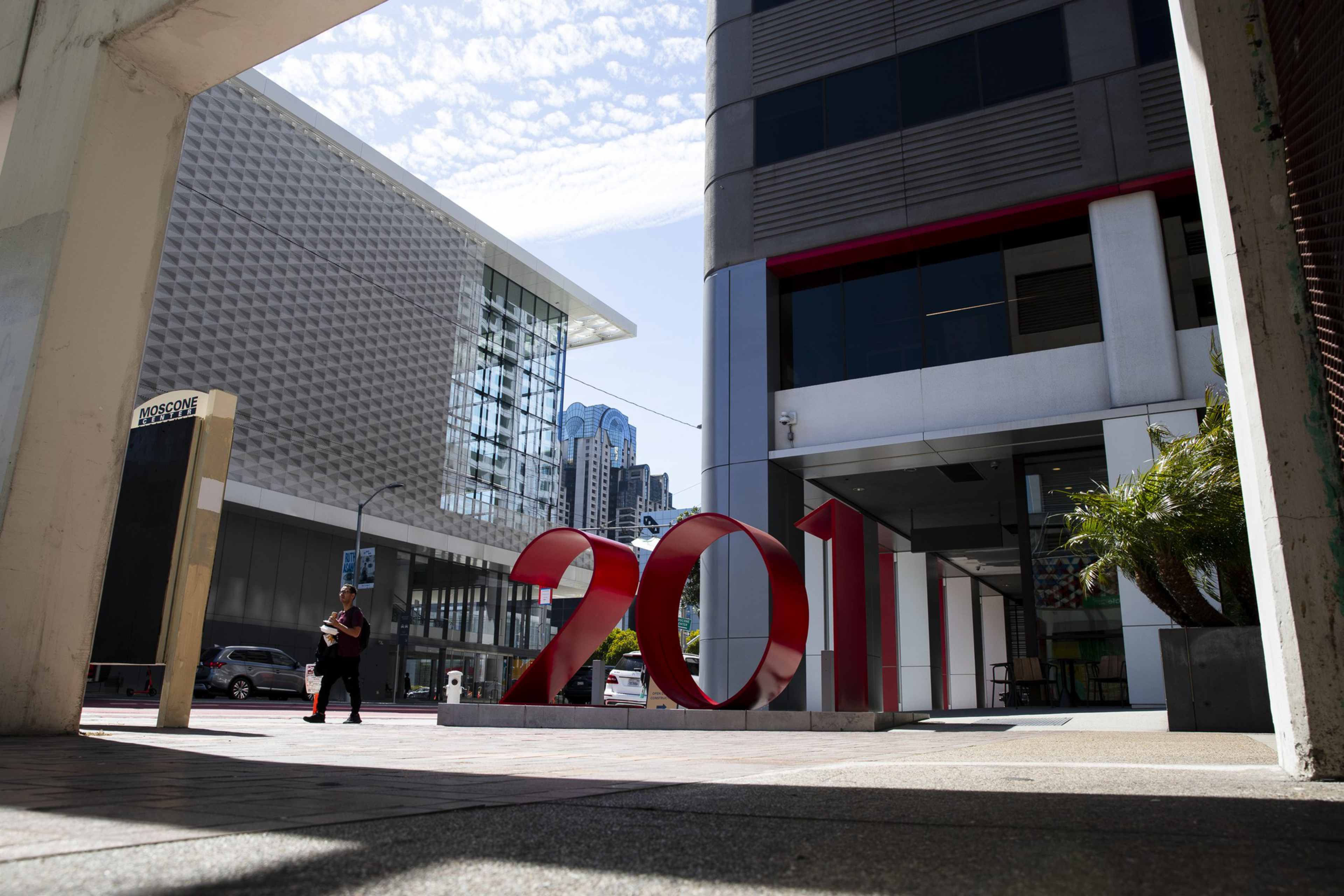 A large red "2011" sculpture stands outside the Moscone Center in a cityscape with modern buildings and a person walking nearby.