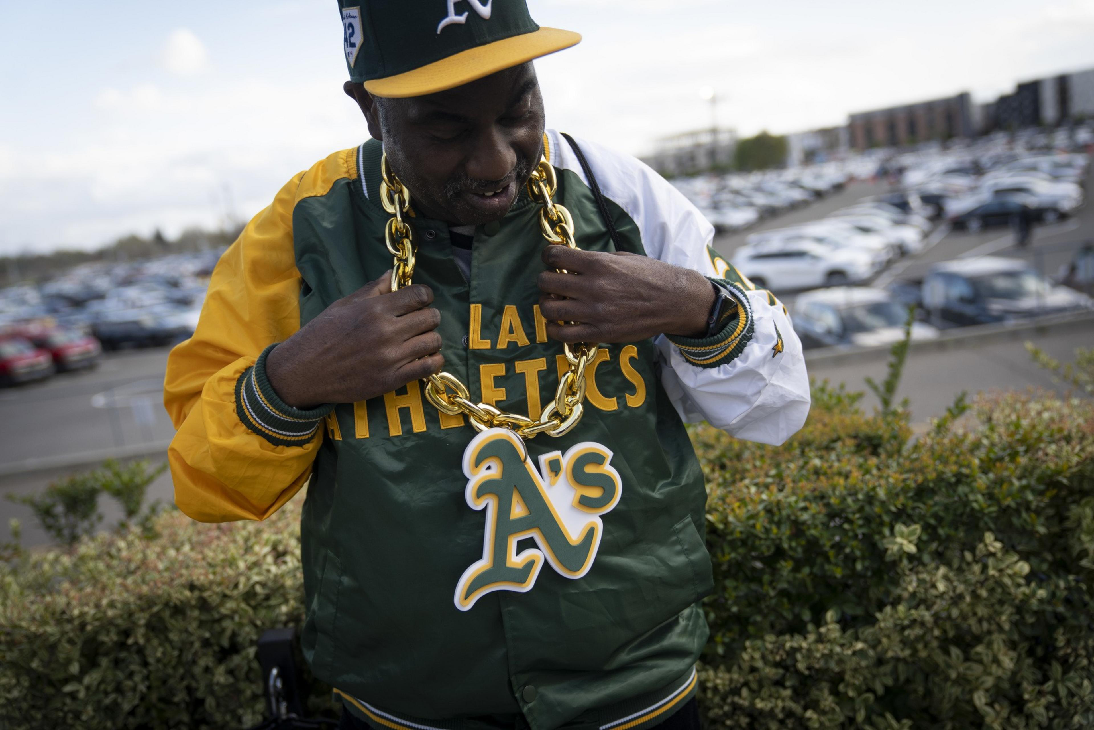 A man in an Oakland Athletics jacket and cap wears a large gold chain with the team's logo. He's outdoors with a parking lot in the background.