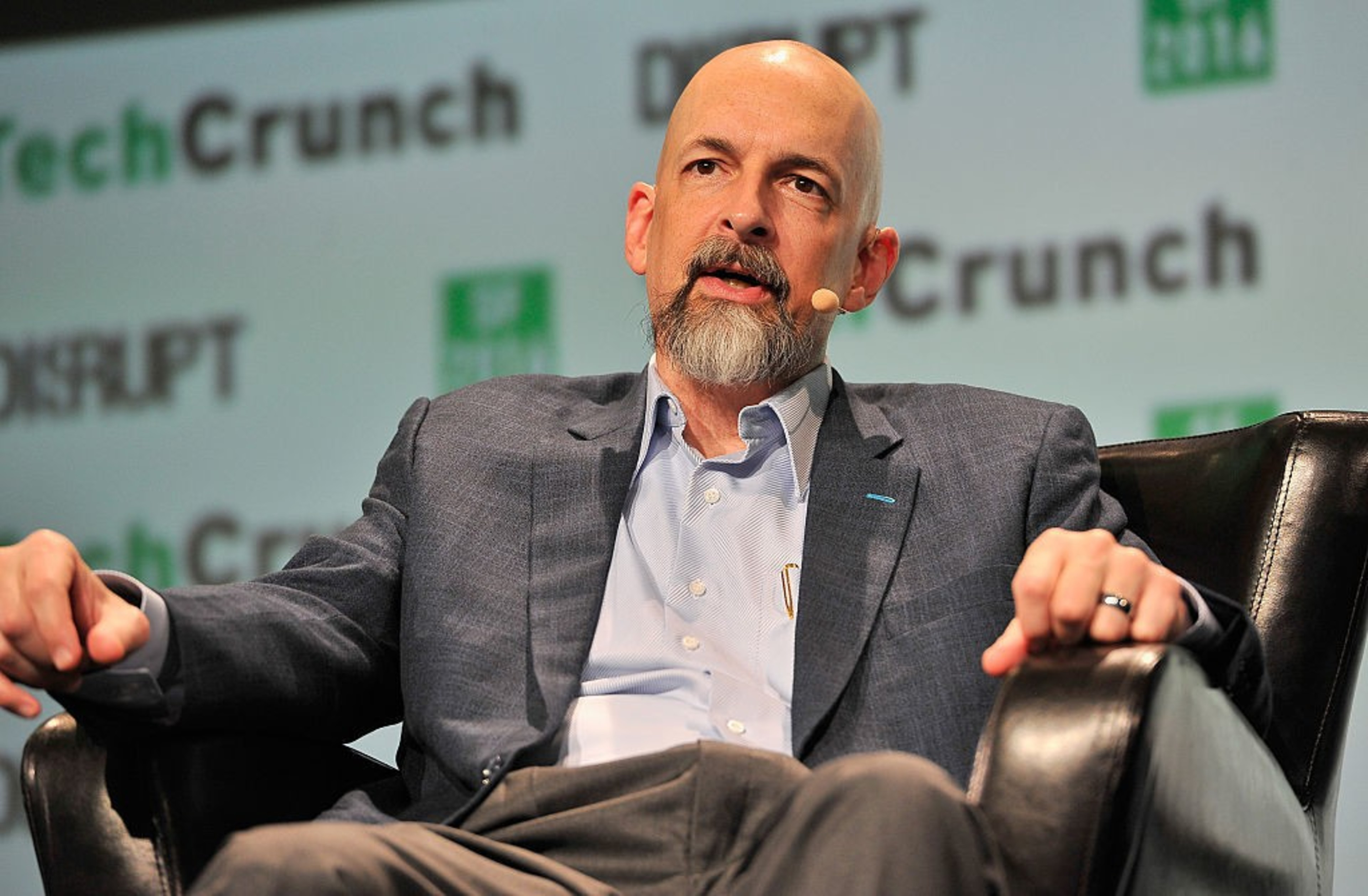 A bald man with a gray beard is speaking at a TechCrunch event, wearing a gray suit and seated in a black chair, holding a microphone near his mouth.