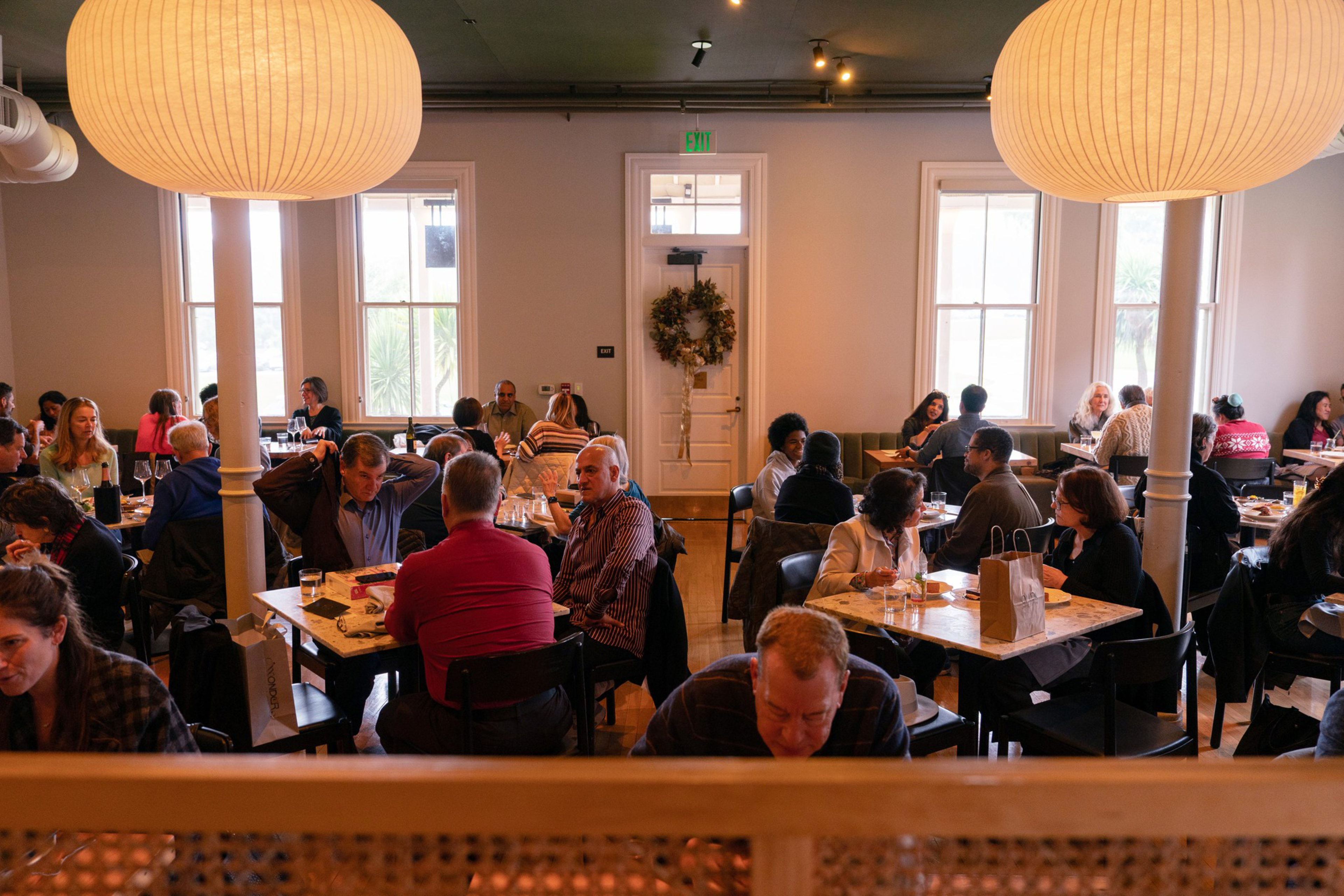 A cozy restaurant with large round lights hangs over patrons dining at tables. The room is filled with people in conversation, creating a lively atmosphere.