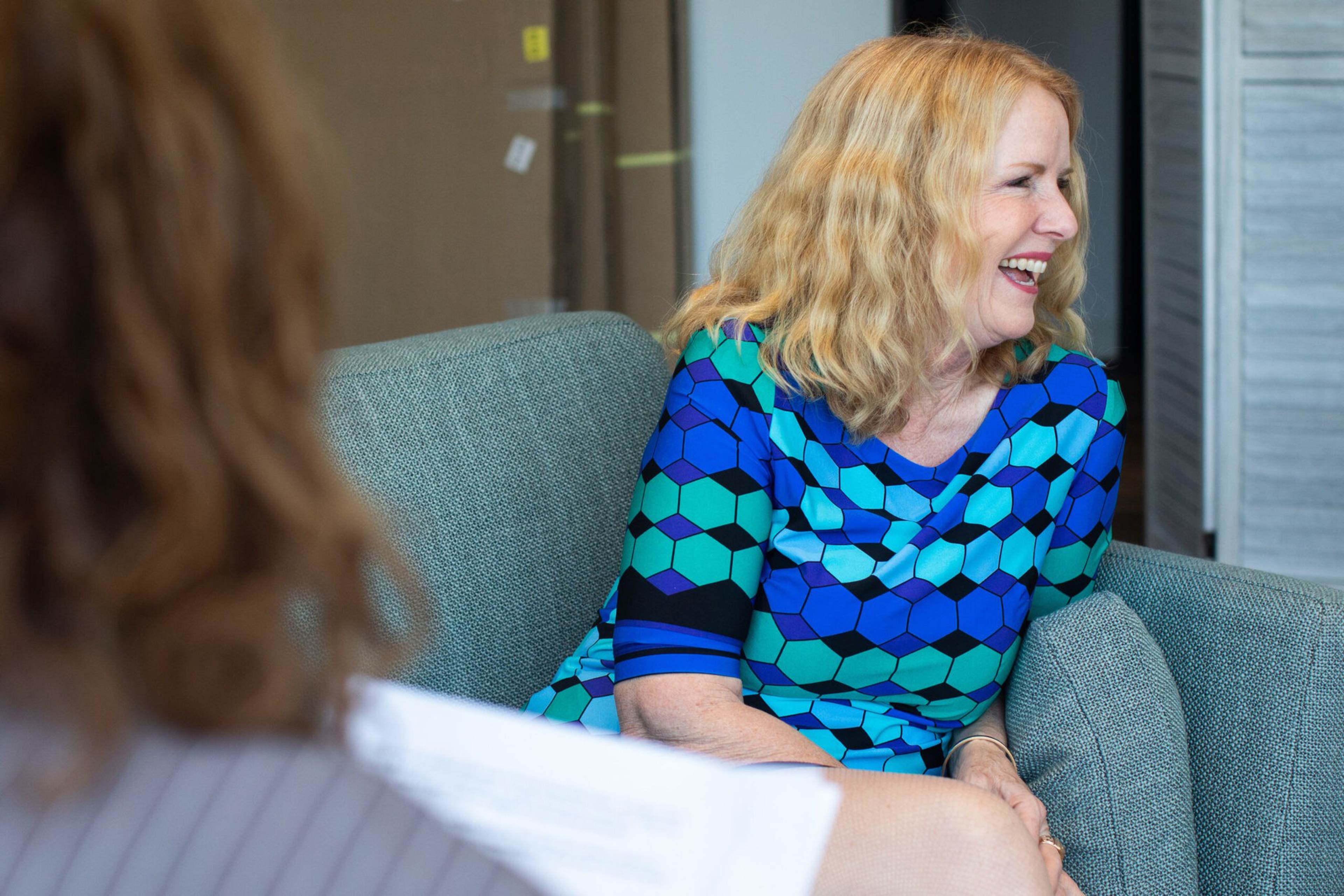 A woman with wavy blonde hair, wearing a blue hexagonal-patterned dress, sits on a gray sofa, smiling and engaging with someone holding papers.