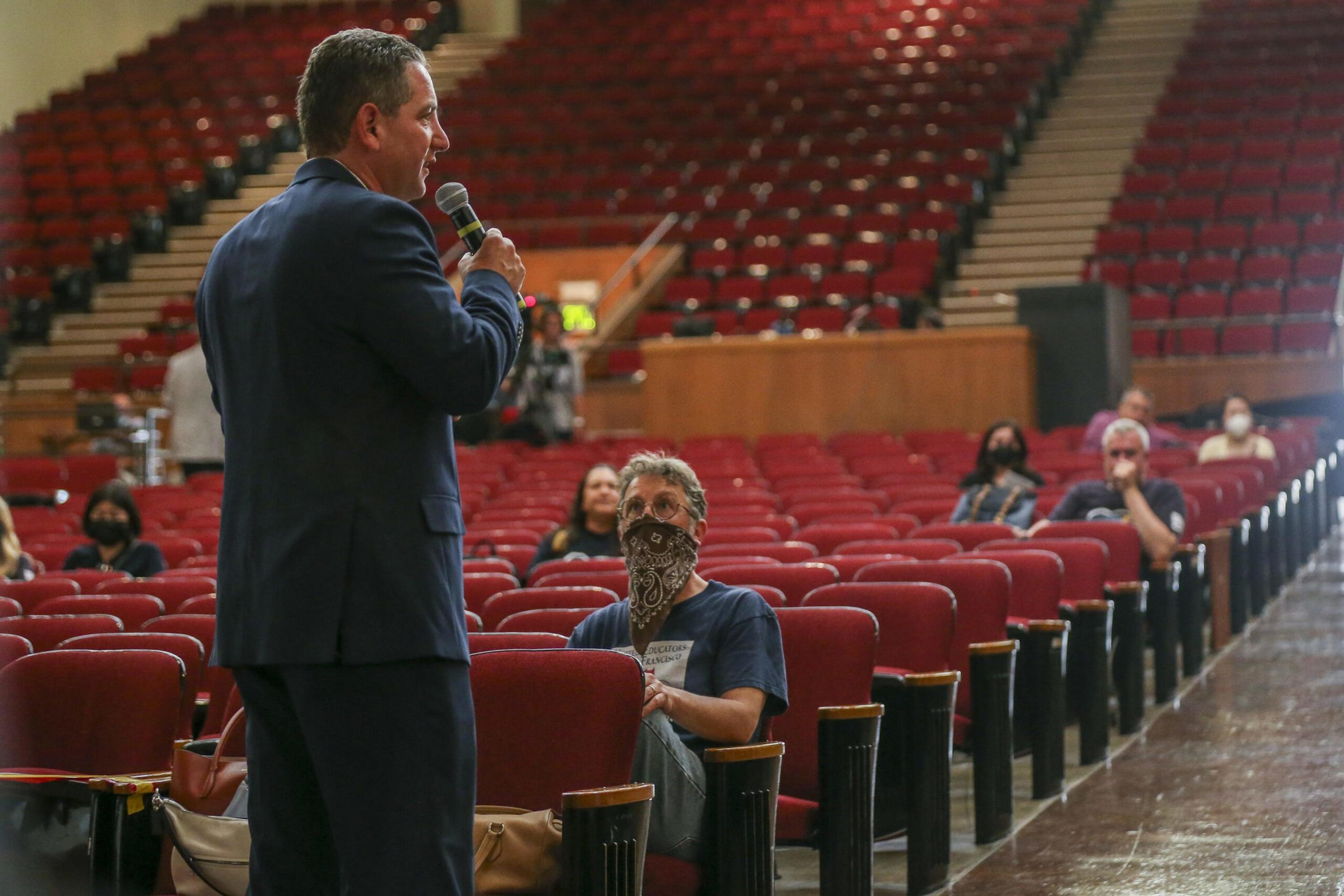 A man in a suit speaks into a microphone facing a small, socially distanced audience wearing masks in a large, mostly empty auditorium.