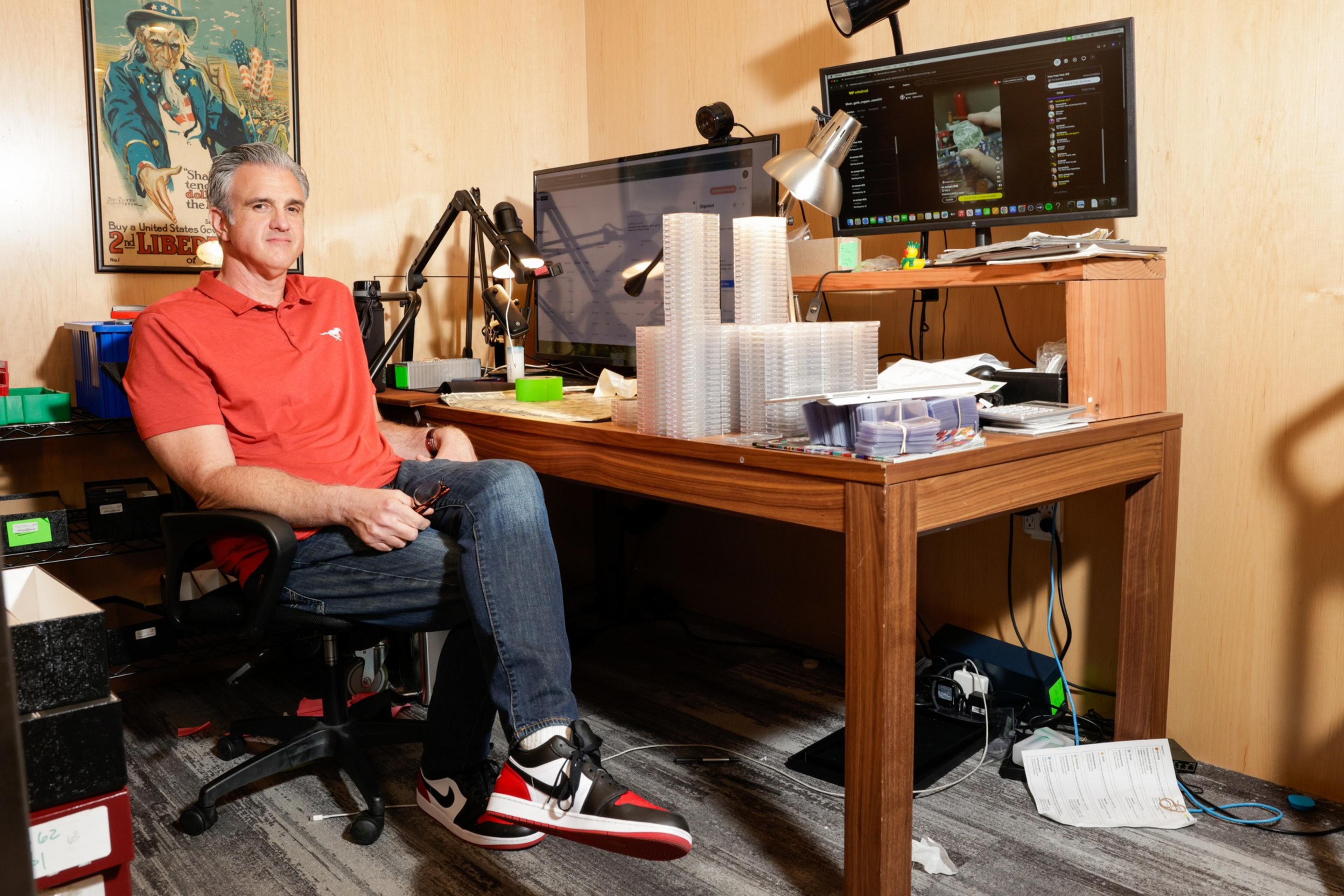 A man in a red shirt and jeans sits in an office, surrounded by stacks of plastic containers and a cluttered desk with monitors and paperwork.
