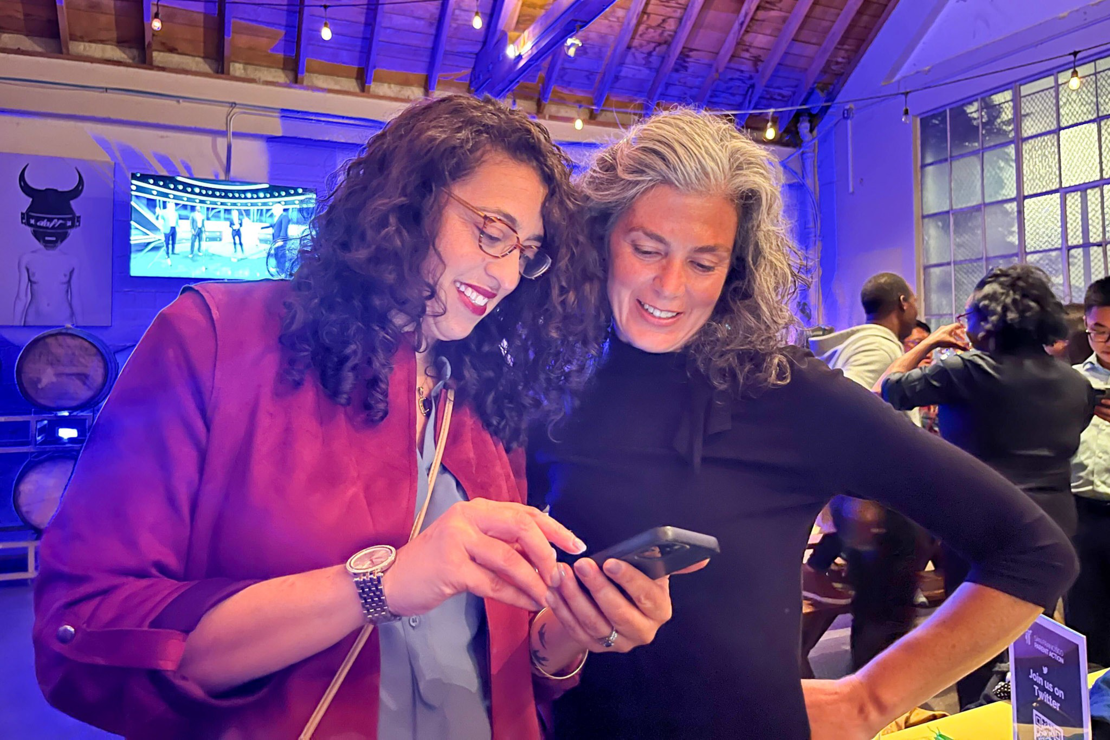 Two women with curly hair smile while looking at a smartphone together at a lively indoor event with purple lighting.