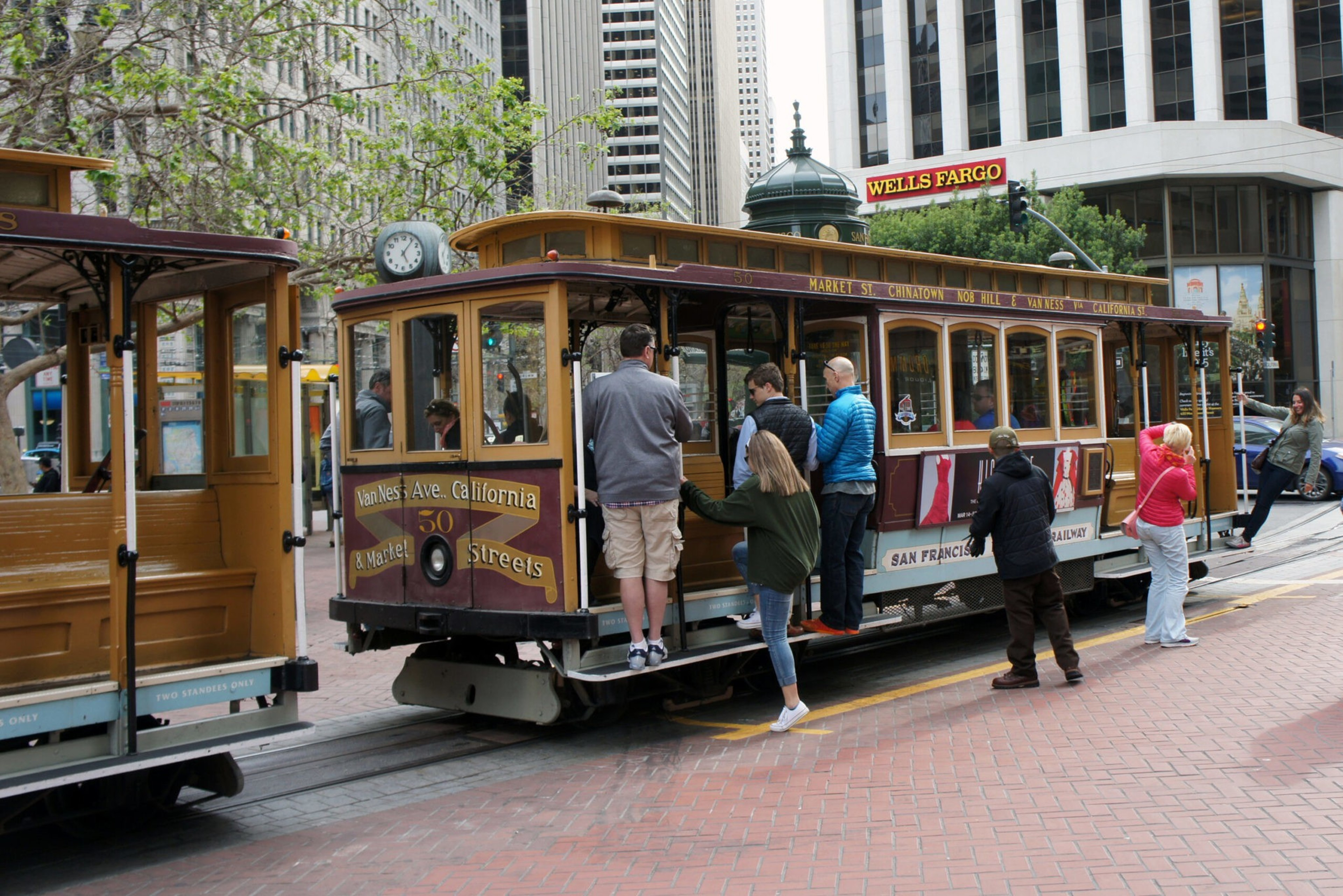 People are boarding a vintage San Francisco cable car at a city stop, with tall buildings and a Wells Fargo sign in the background.