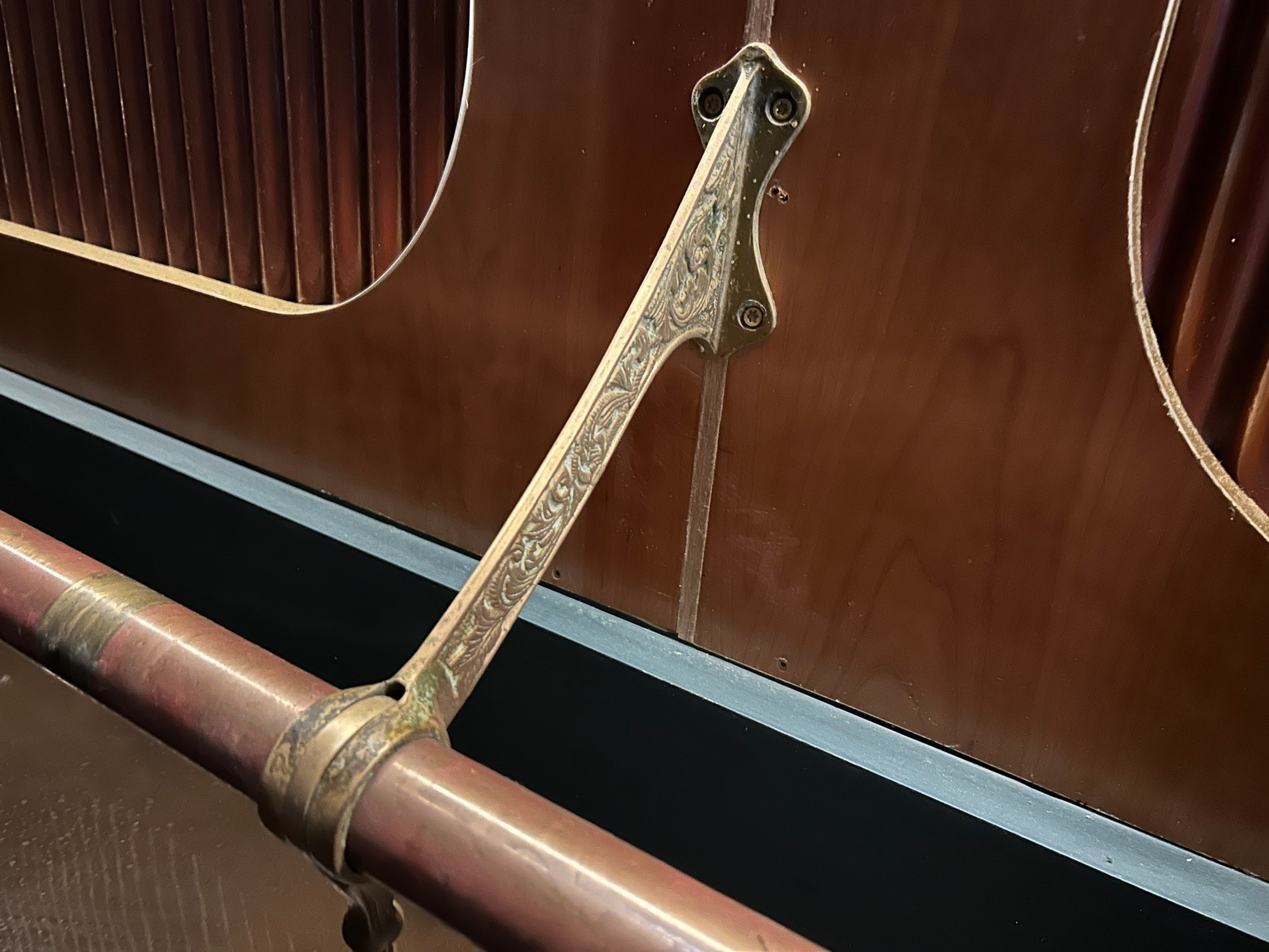 A close-up of an engraved brass sled runner brace attached to a curved wooden sleigh side, with a maroon metal rail running horizontally below it.
