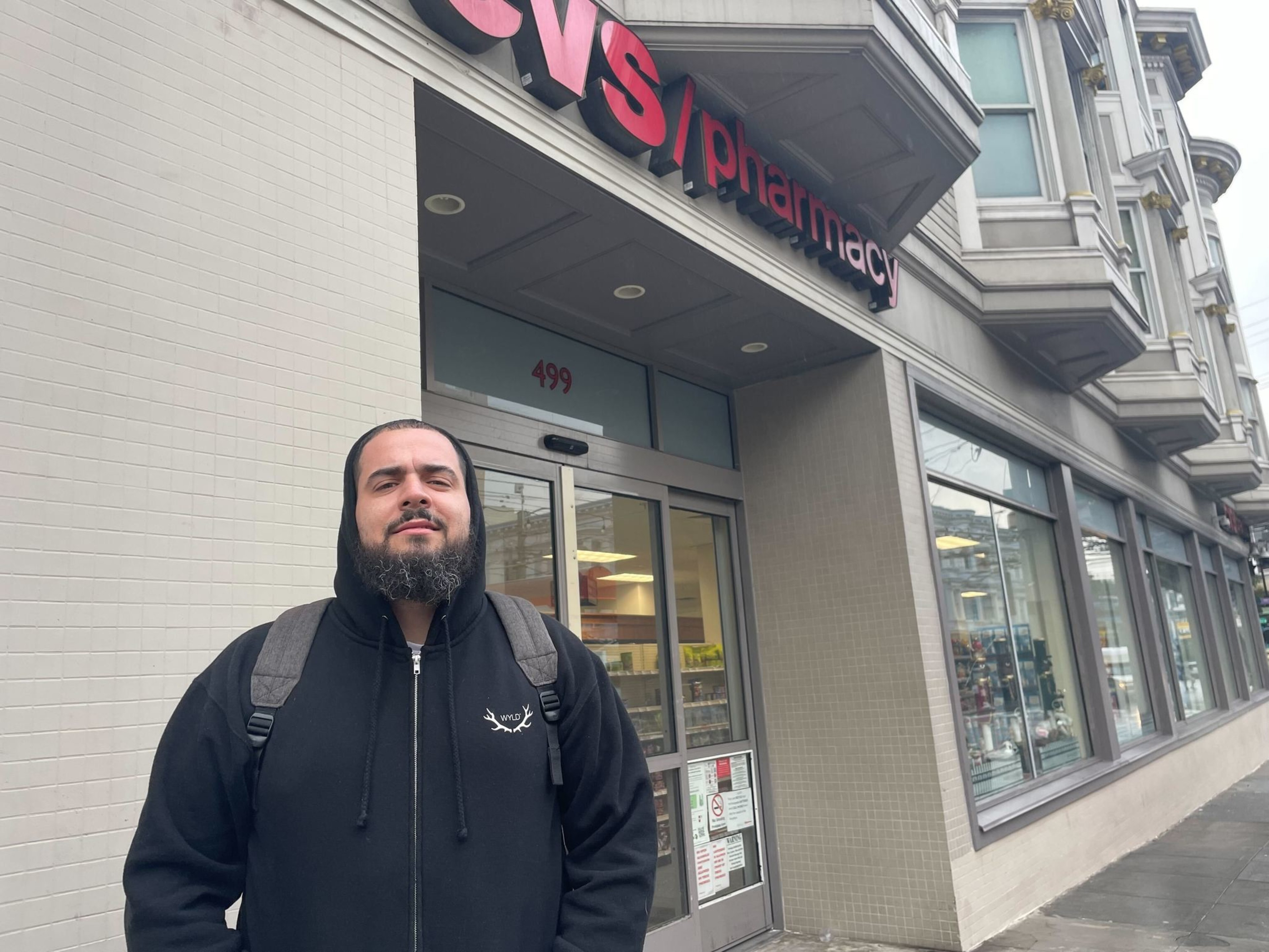 Man in zip-up hoodie wearing backpack stands on sidewalk outside CVS store