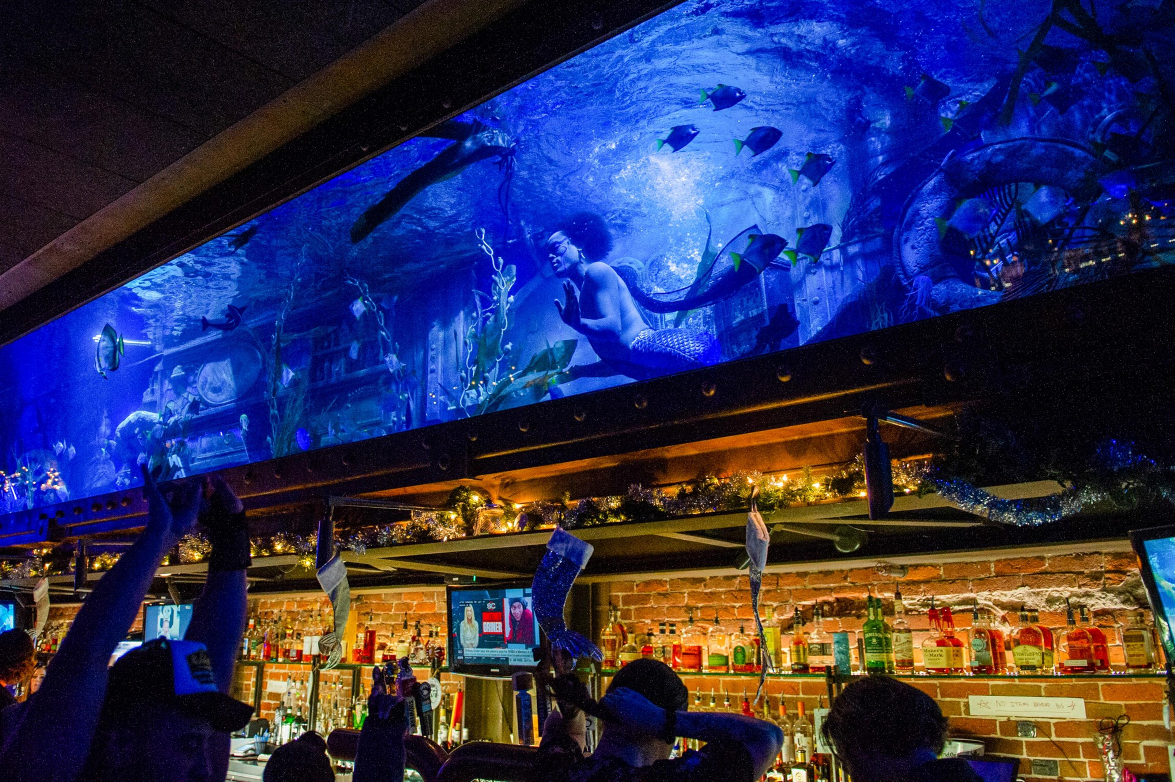 A bar with people raising glasses under a large, illuminated aquarium featuring a person posing underwater like a mermaid, surrounded by fish and underwater plants.