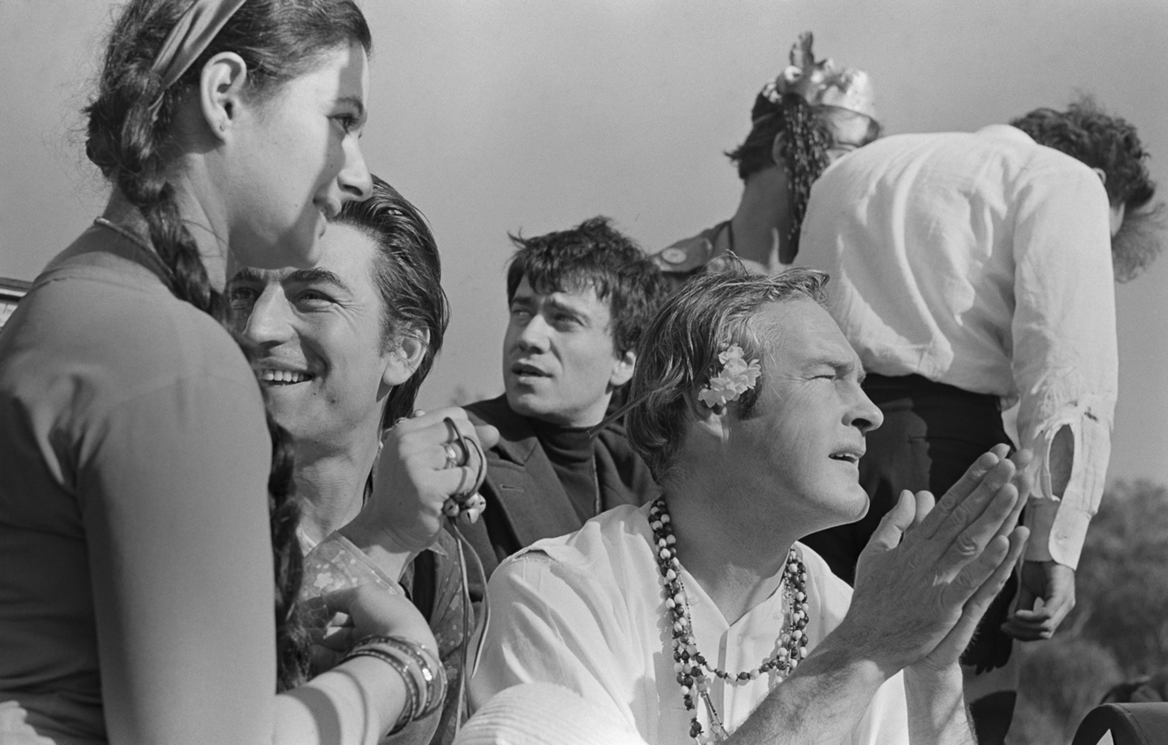 A group of people appears to be enjoying an outdoor gathering. One man with a flower in his hair clasps his hands, while others engage and smile, creating a lively scene.