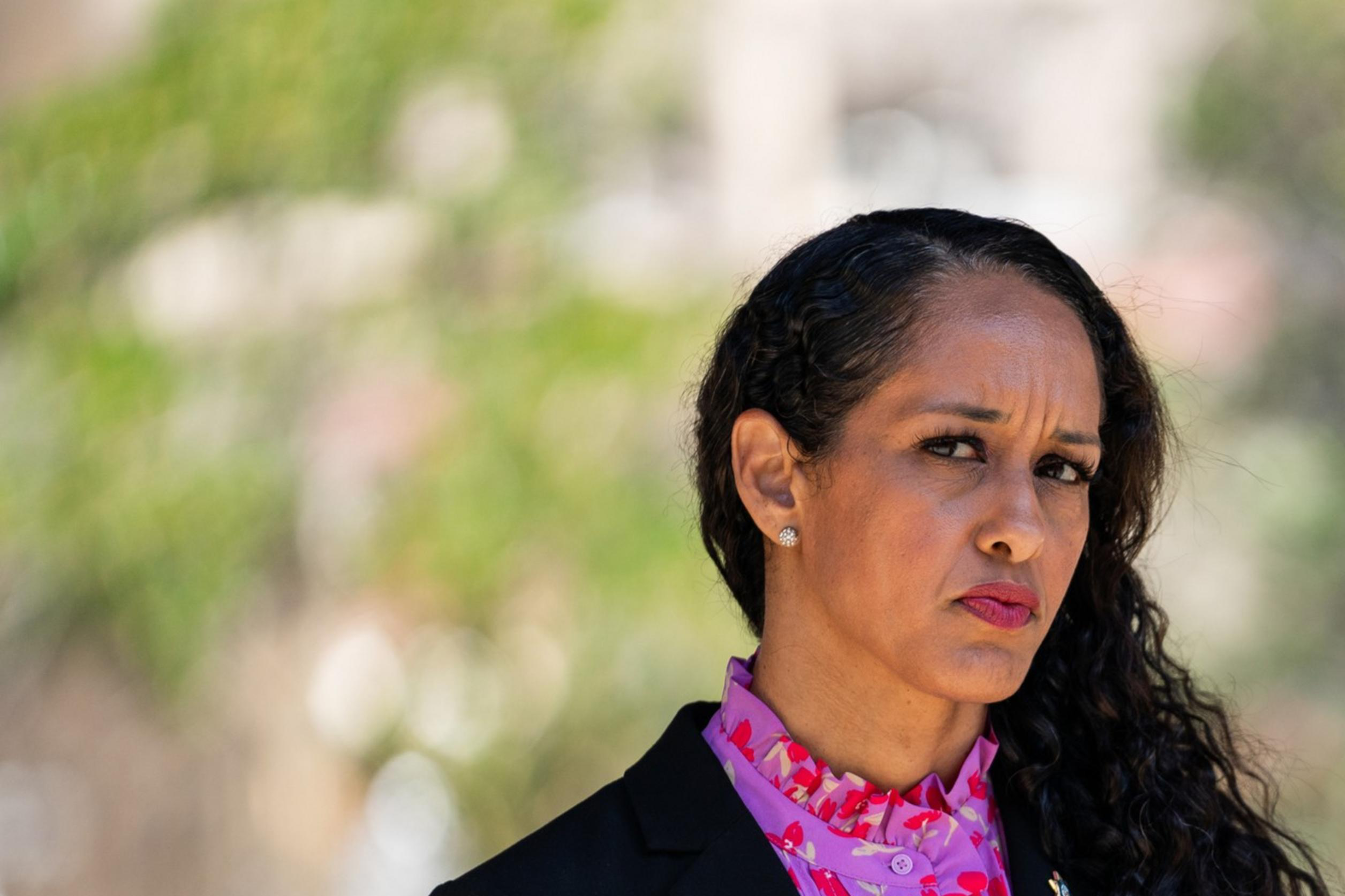 A woman with long, wavy hair in a black blazer and bright pink, floral blouse looks forward with a serious expression. The background is blurred and green.