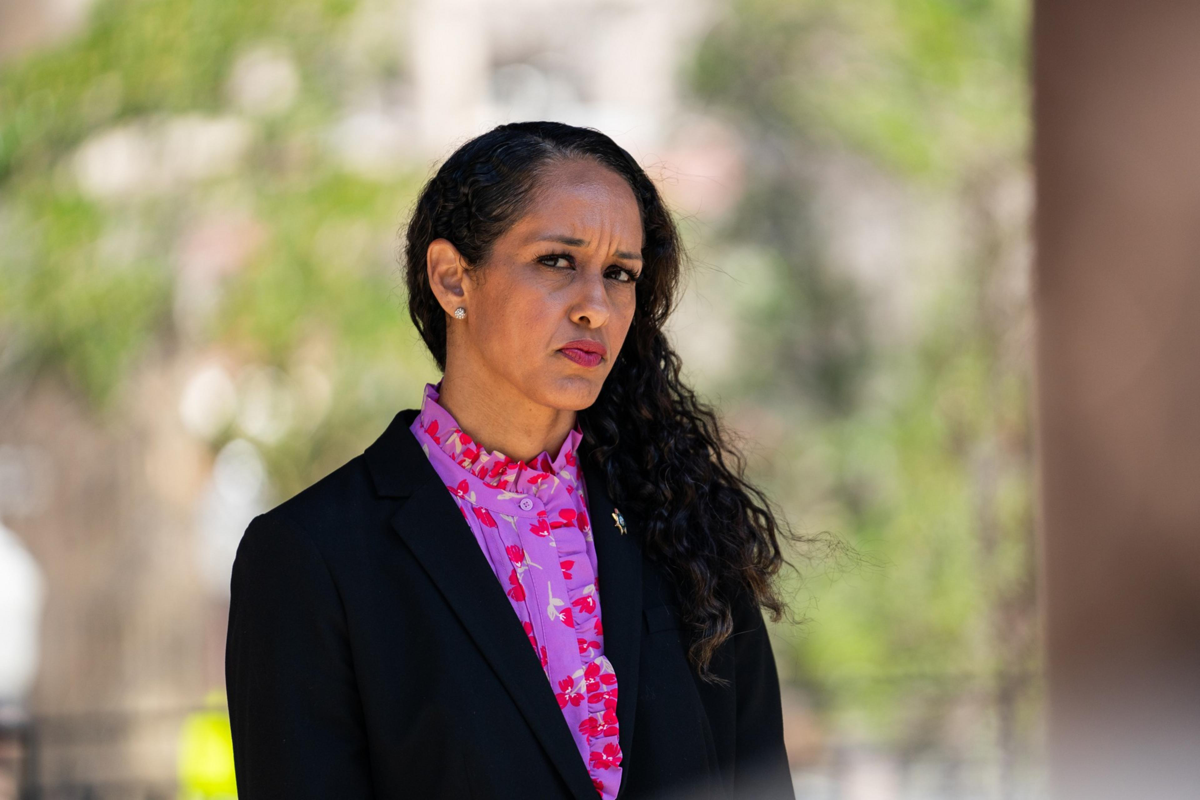 A woman with long, wavy hair in a black blazer and bright pink, floral blouse looks forward with a serious expression. The background is blurred and green.
