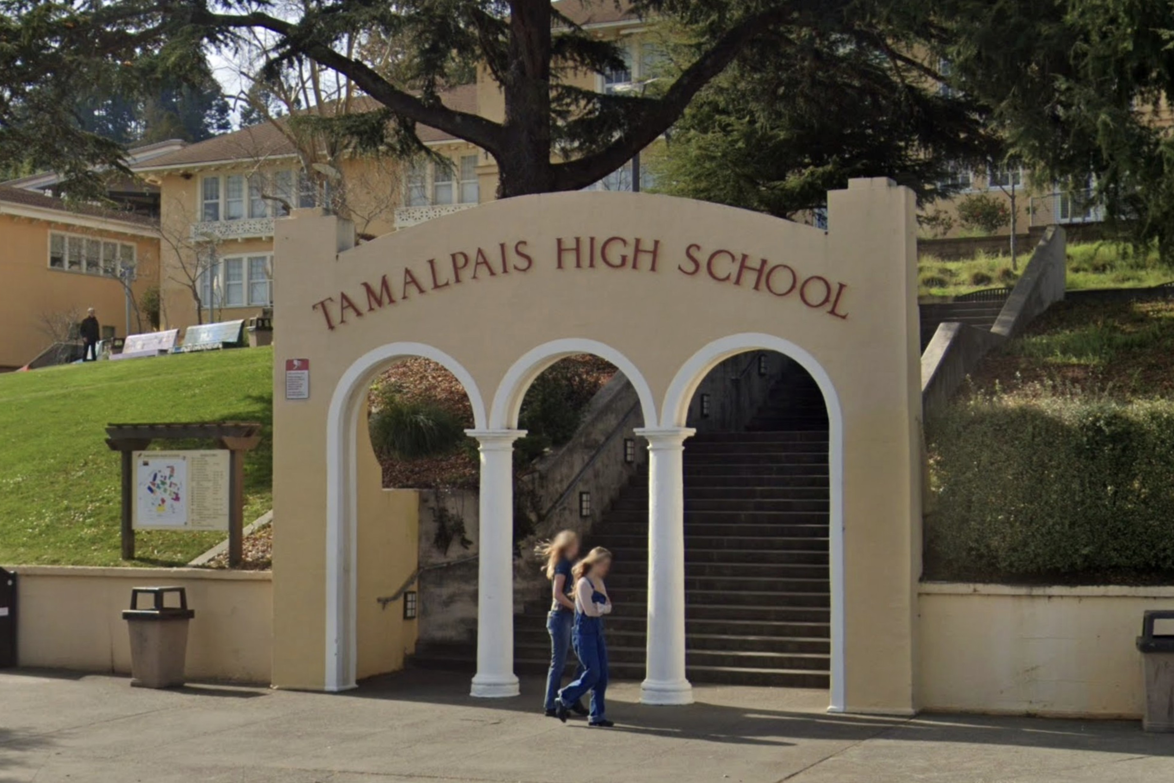 The image shows a beige archway with "Tamalpais High School" written on it, at the base of stairs leading up to a building surrounded by trees and greenery. Two people walk by.