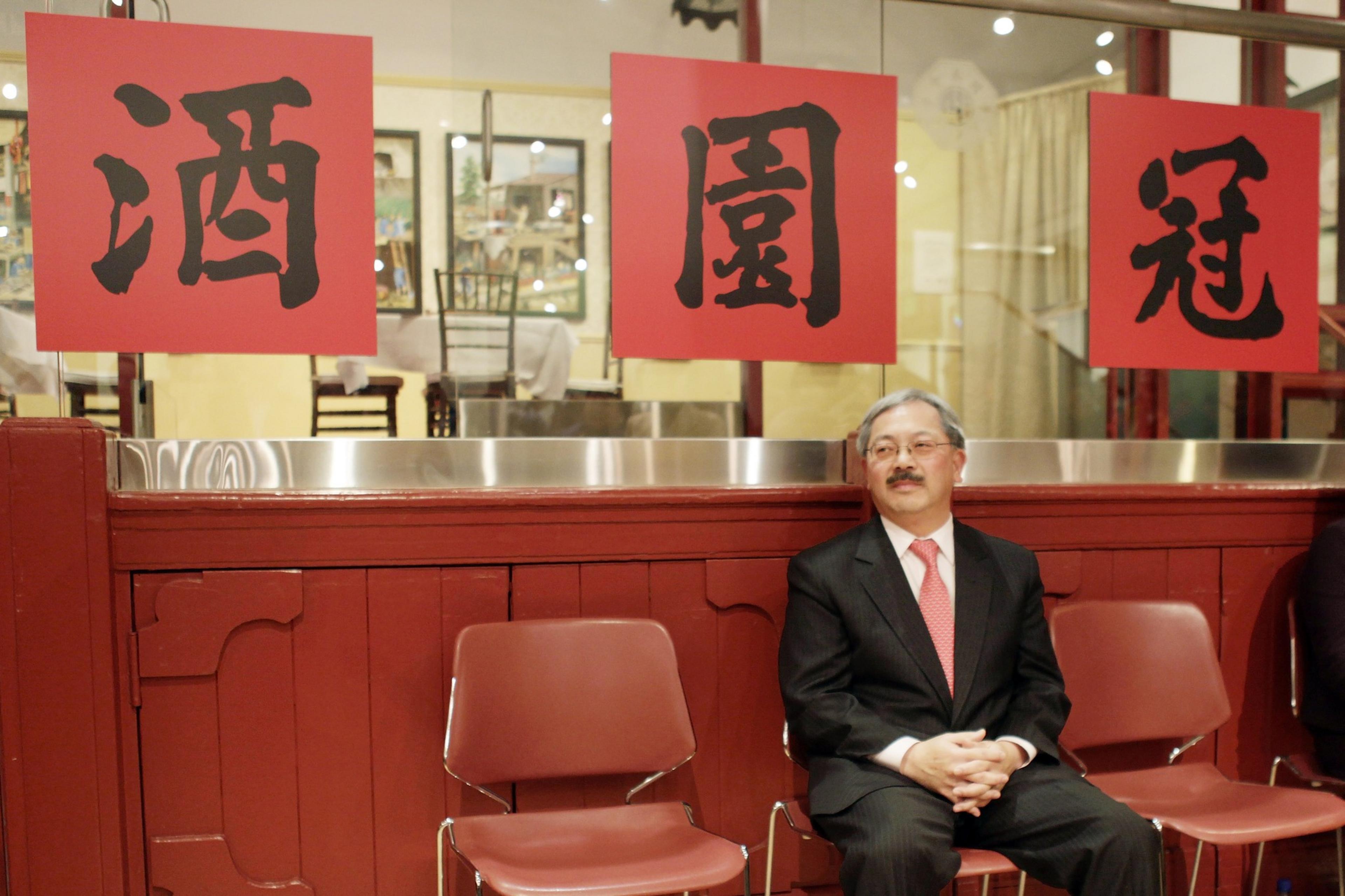 A man in a black suit and pink tie sits with folded hands between two empty red chairs under three red signs with black Chinese characters.