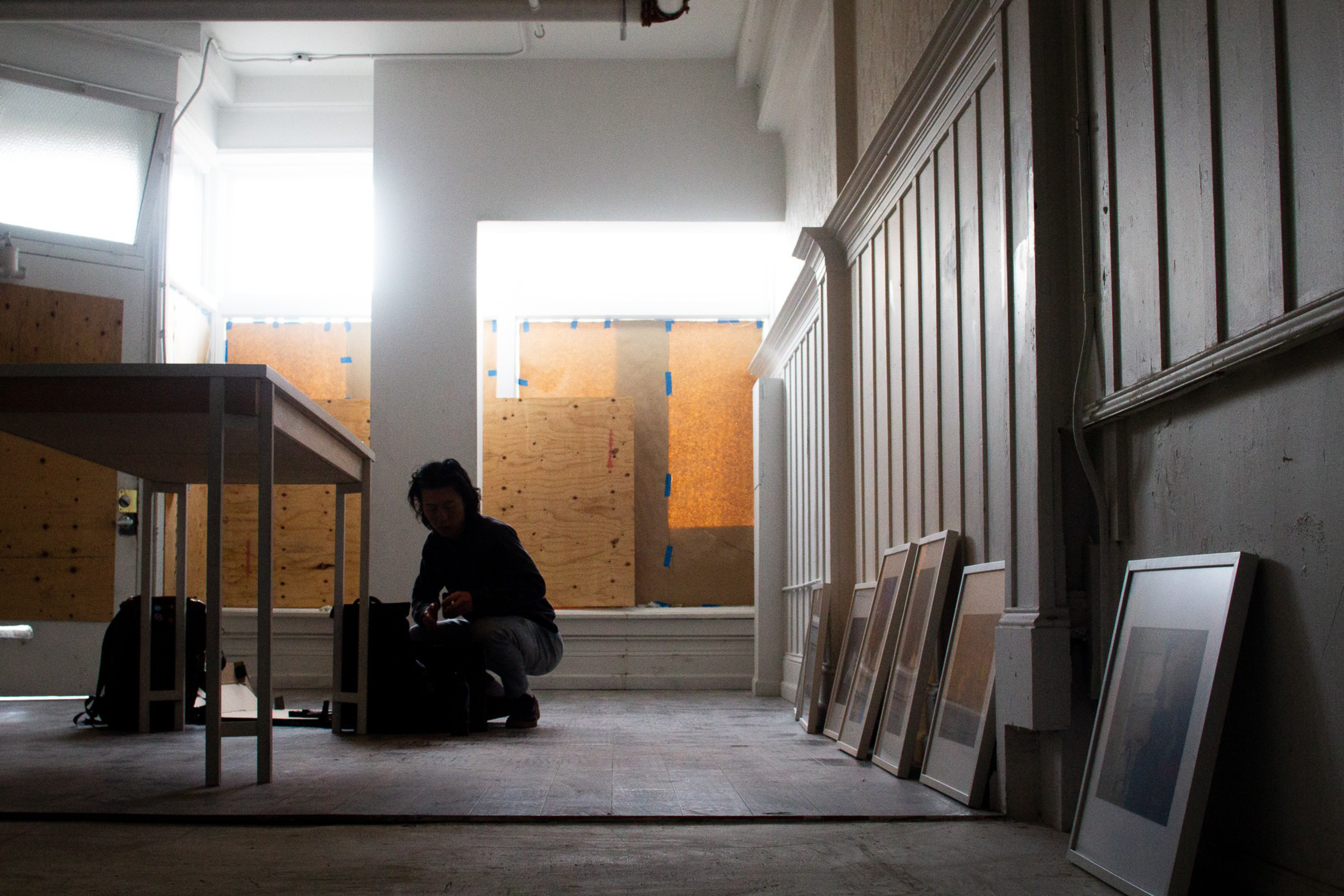 A dimly lit room with boarded-up windows features a person crouching near a table and several framed pictures leaning against a wall.