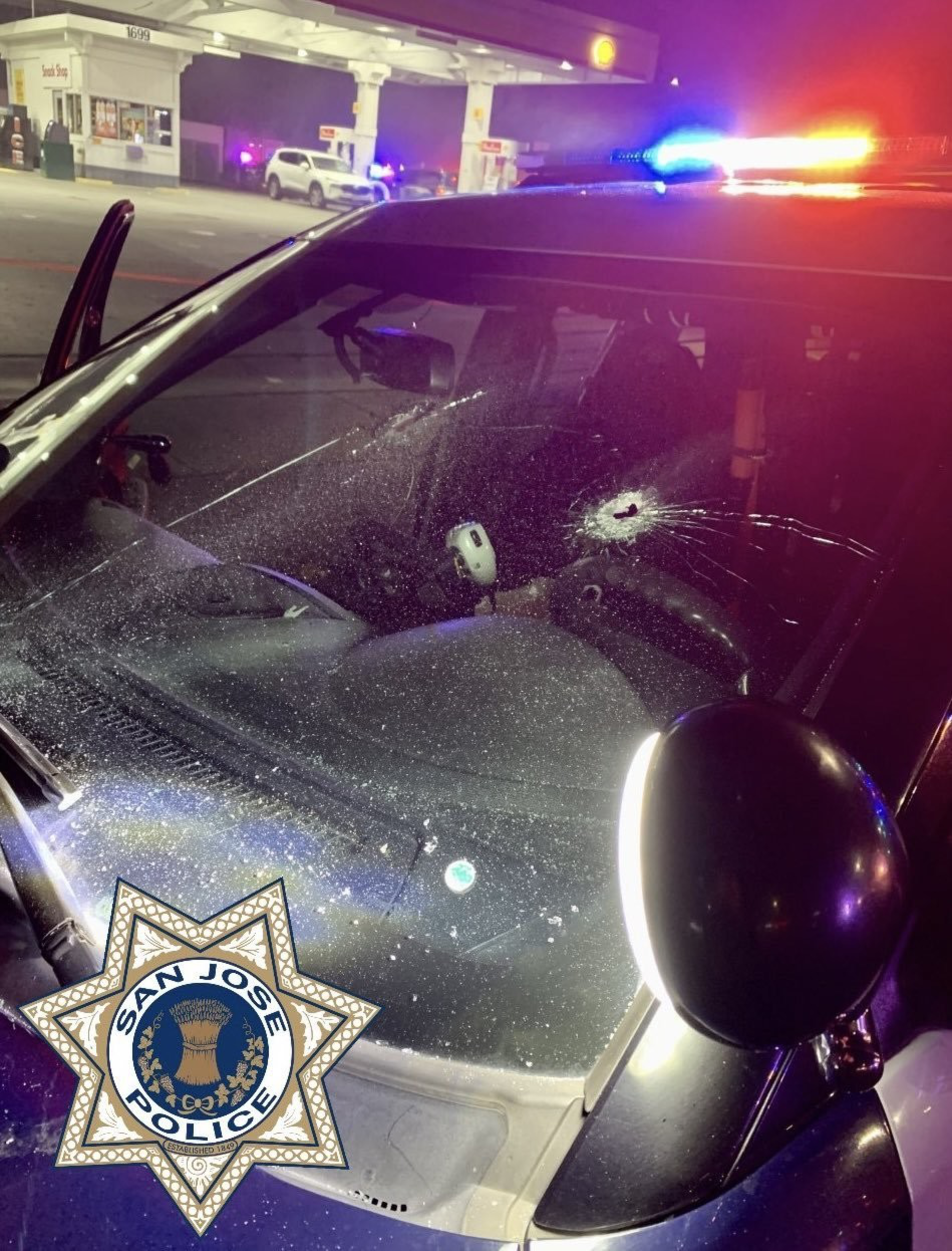 A police car with flashing red and blue lights has a bullet hole in its windshield, with a shattered area radiating from the impact point near the driver's seat.