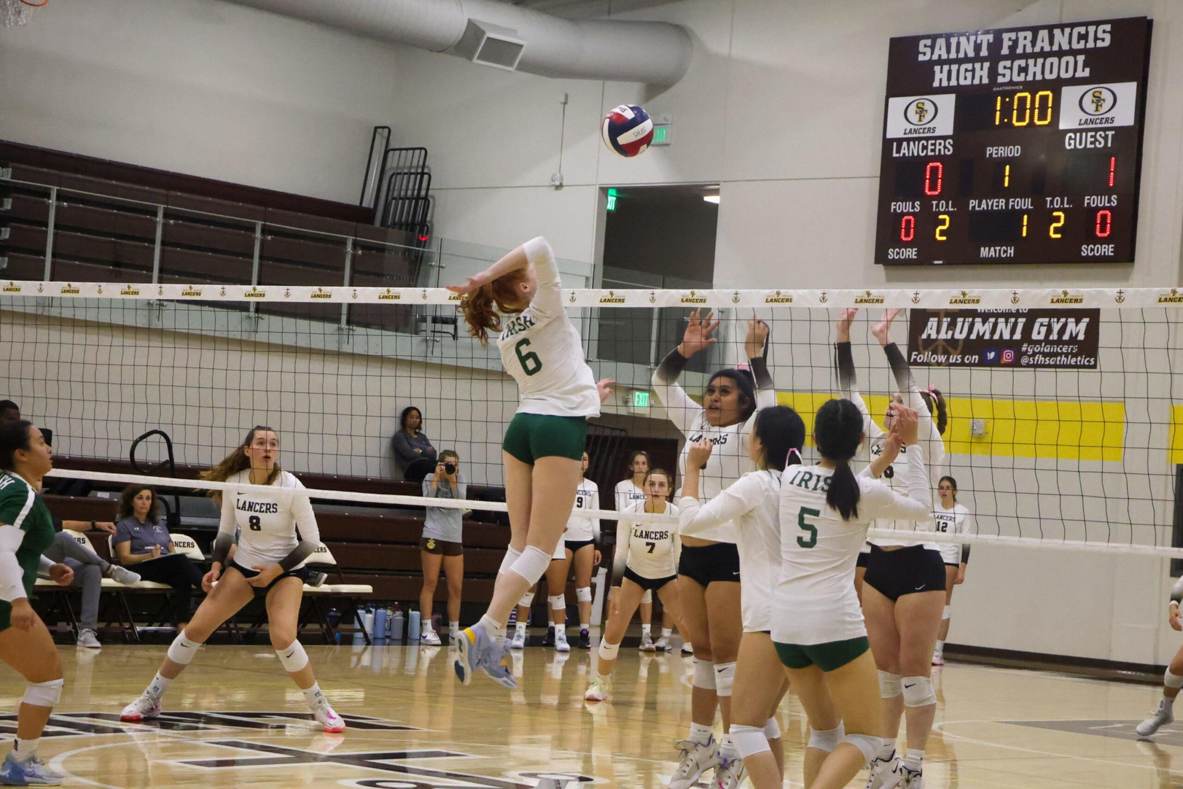 A volleyball player in a white and green uniform jumps to hit the ball over the net while opposing players in white and black uniforms raise their hands to block.