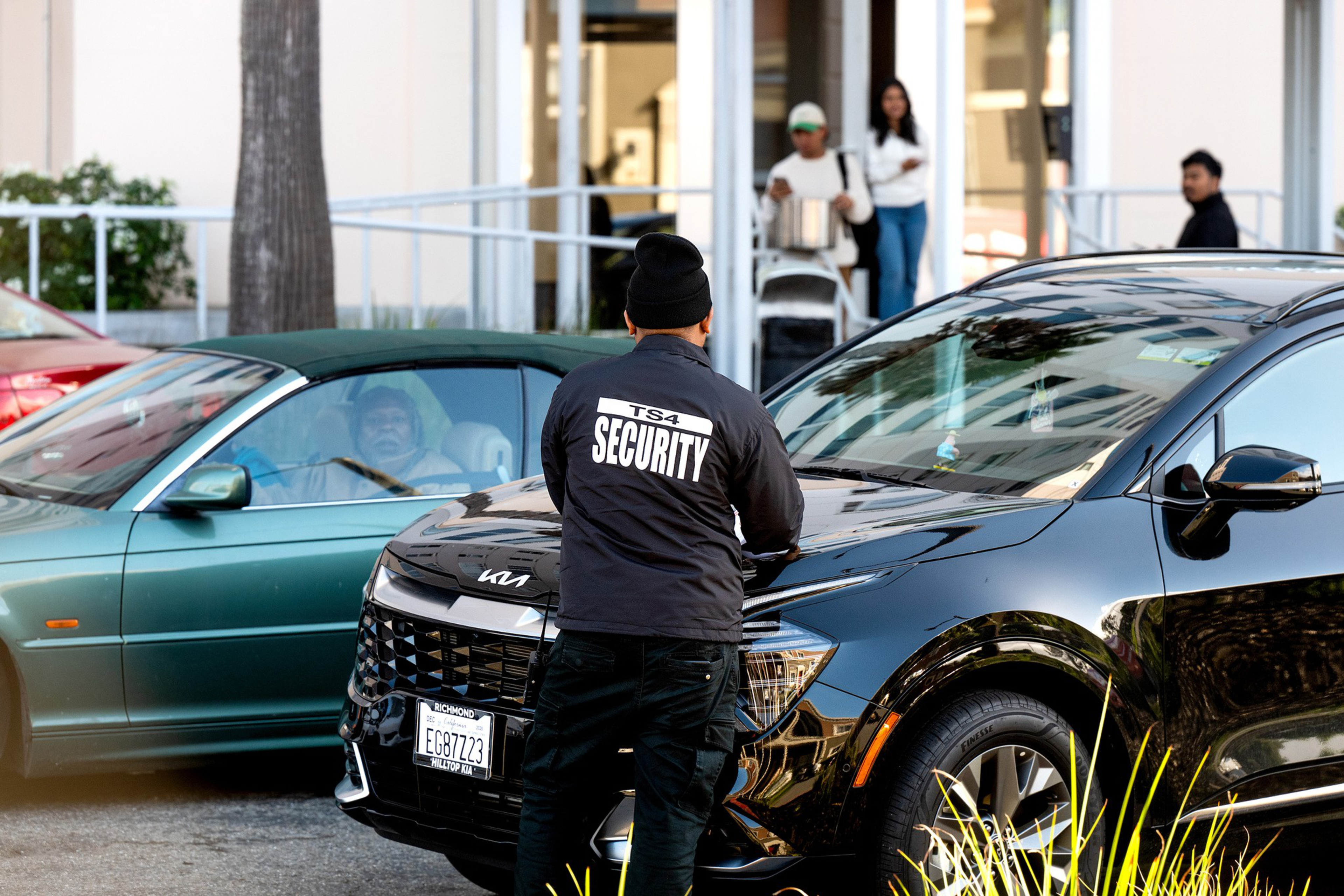 A man in a black “Security” jacket stands next to a black Kia SUV, while a person in a green car watches and three people stand on the sidewalk behind them.