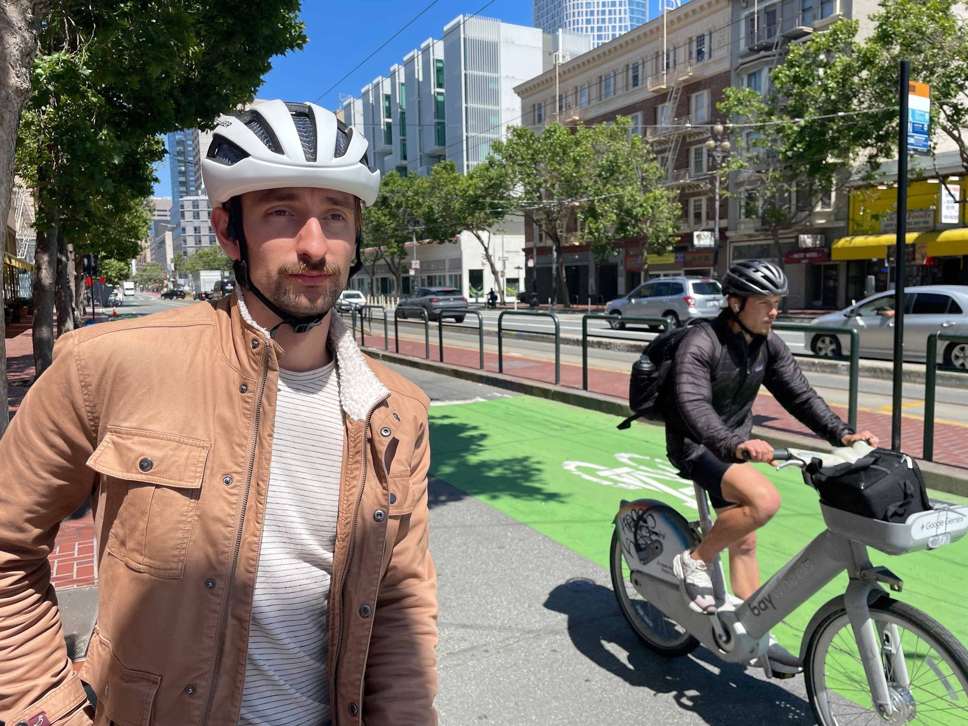 A man in a tan jacket and white helmet stands by a green bike lane while another man rides a gray bike wearing a black helmet and jacket.