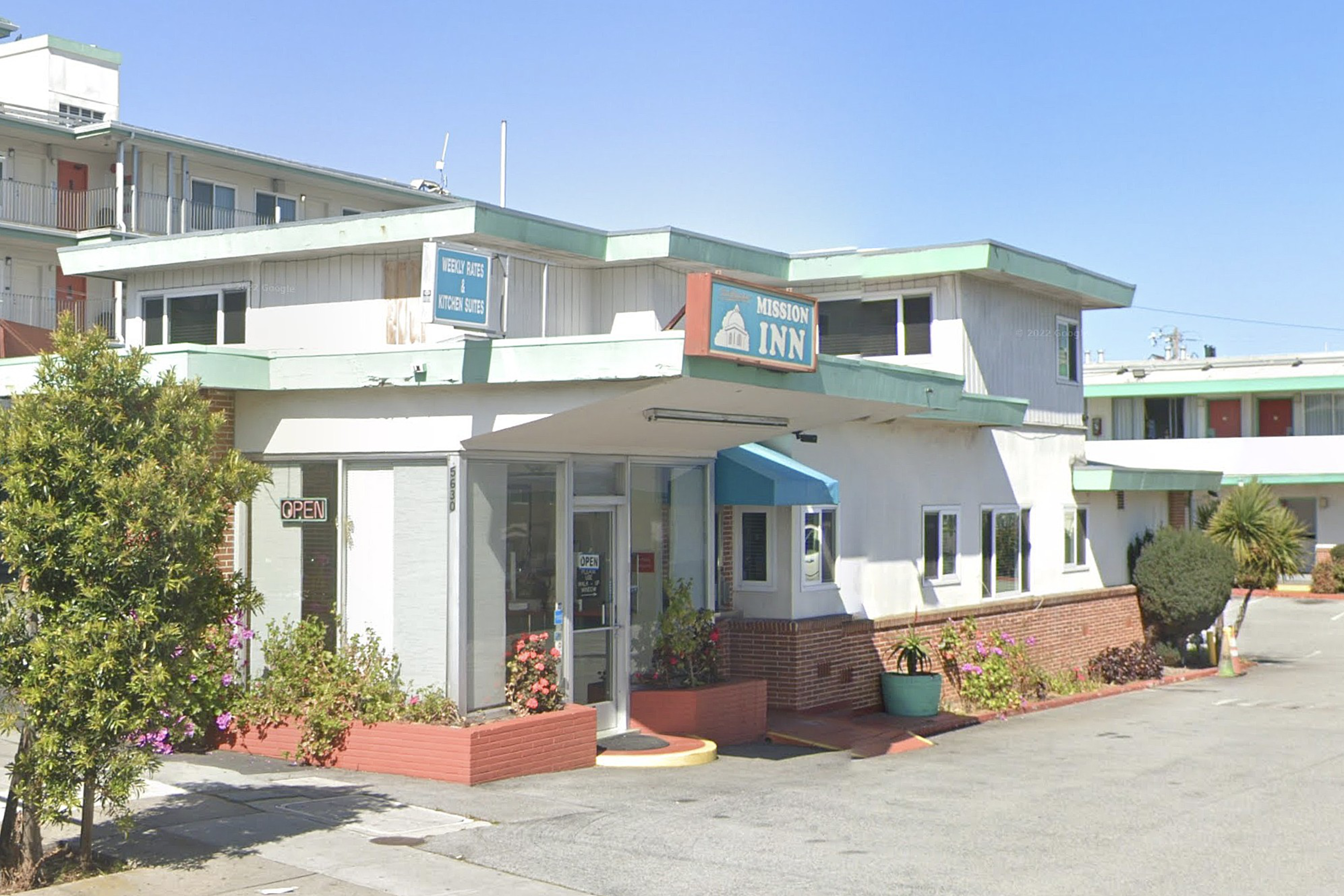 A two-story motel named Mission Inn with white walls, green roof edges, plants around the entrance, and a clear blue sky above.