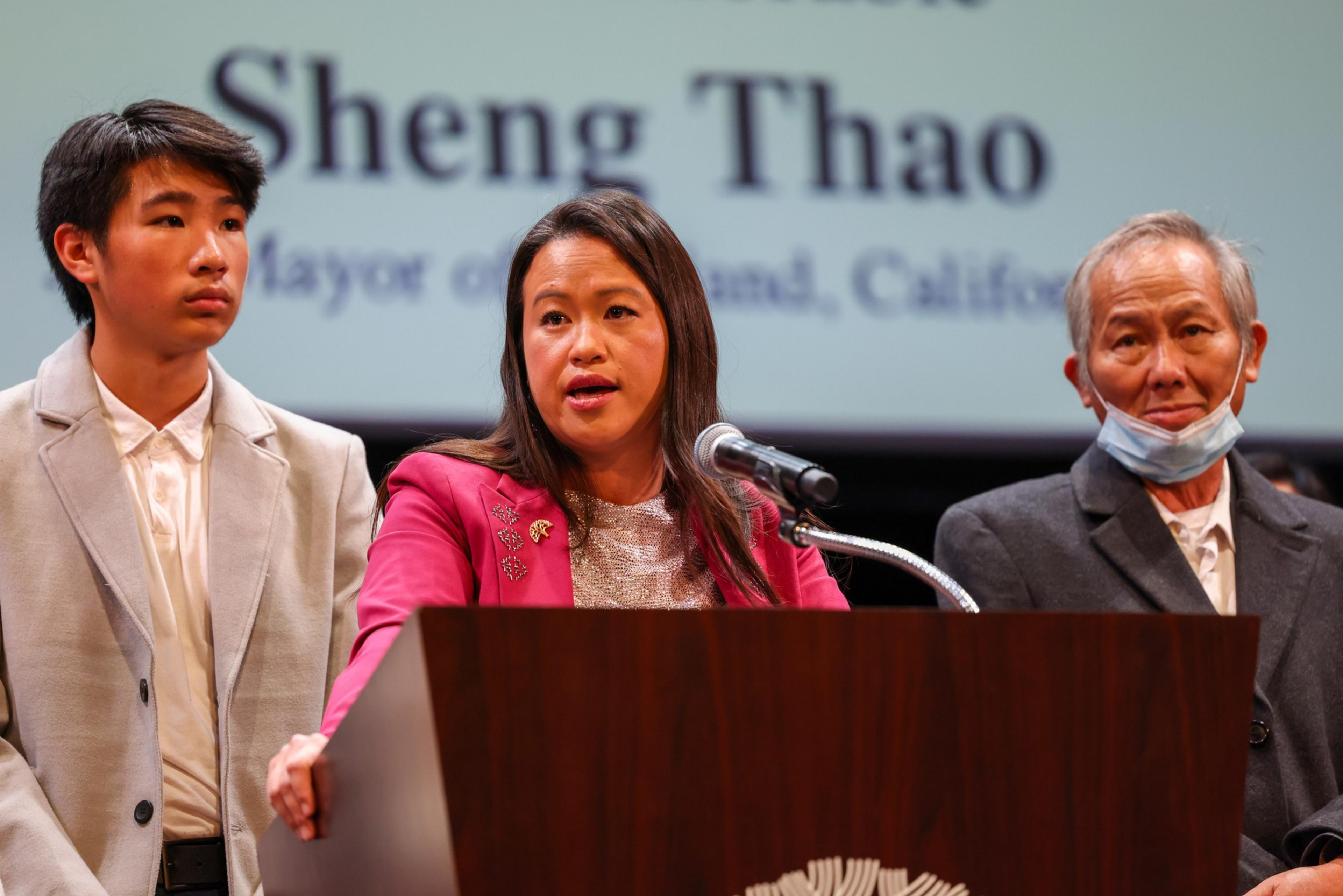 A woman in a pink jacket speaks at a podium, flanked by a young man in a light suit and an older man wearing a partially lowered face mask.