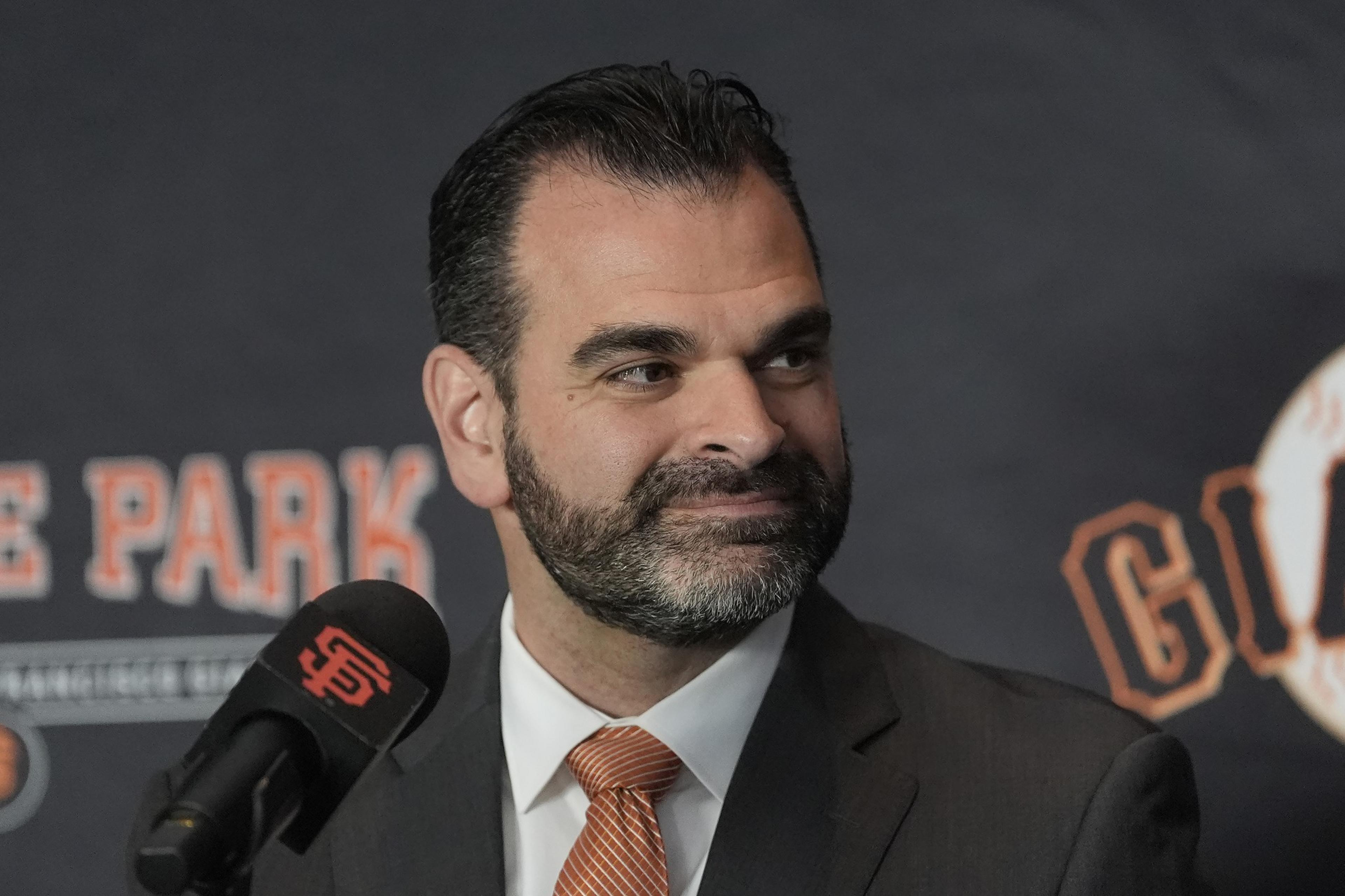 A man in a suit with a beard smiles slightly, standing in front of a backdrop with the words "GIANTS" and "PARK." A microphone is in front of him.