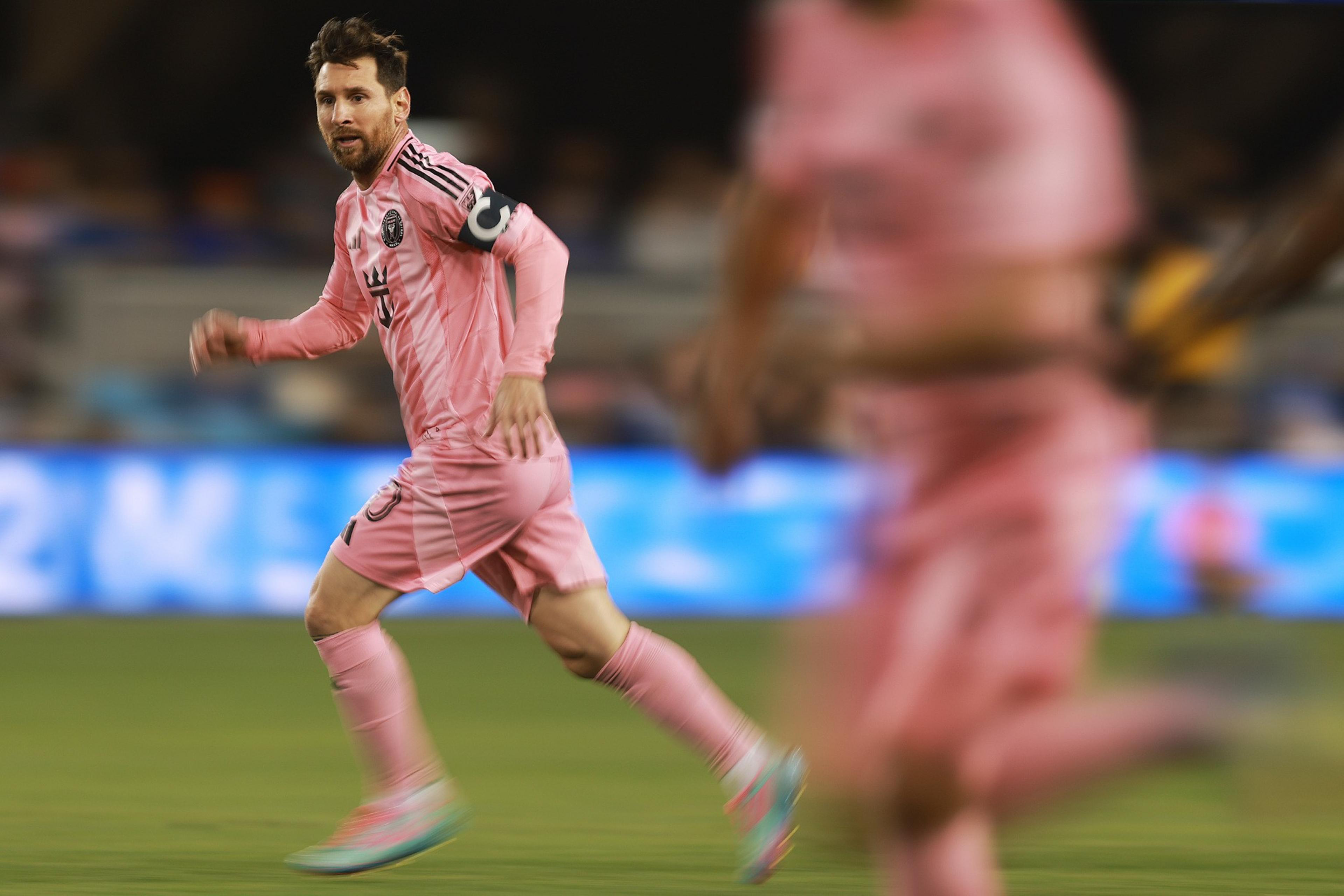 A soccer player in a pink uniform with a captain's armband is running on the field, focused on the game. Another player is partially visible.