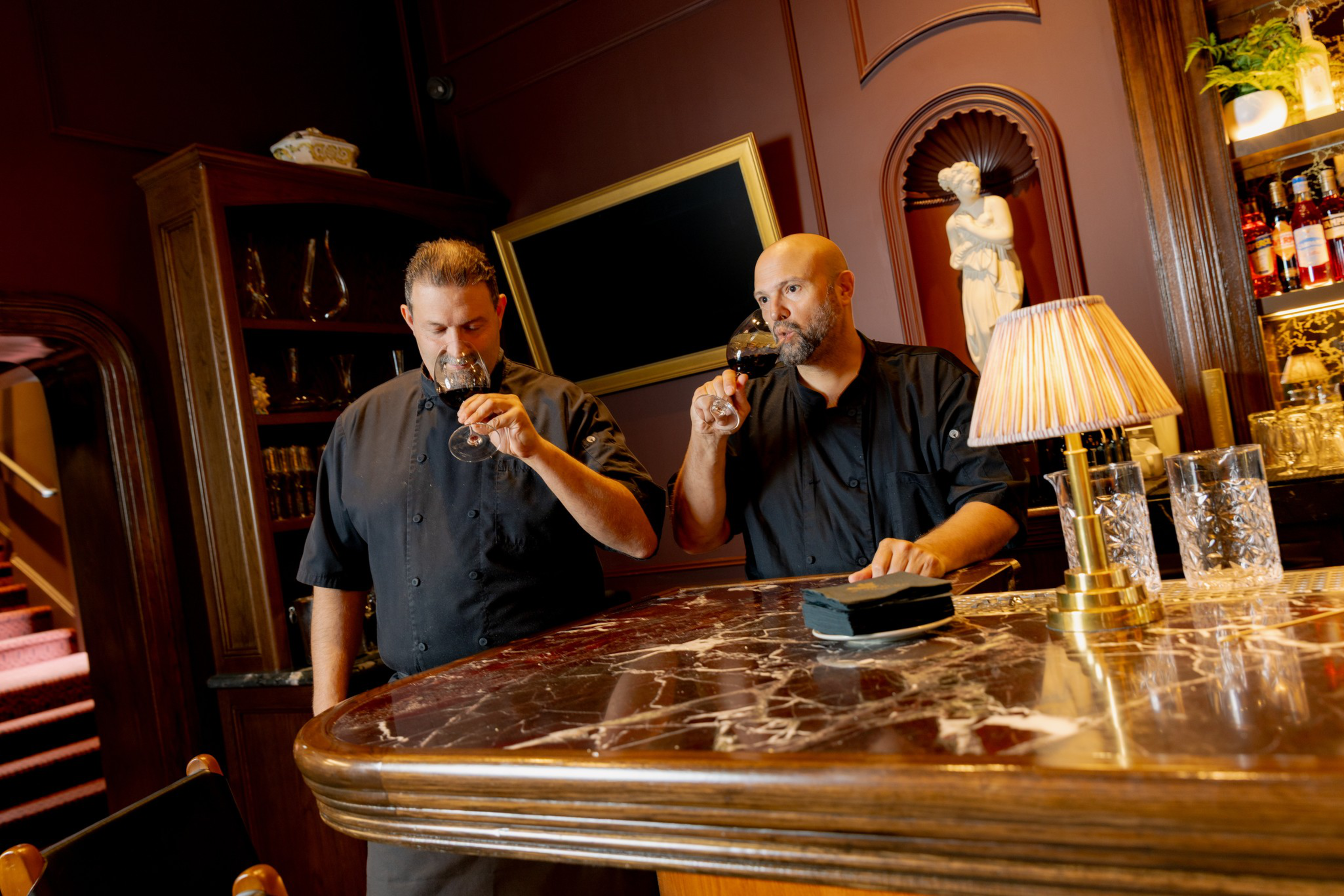 Two men in black shirts sip red wine at a polished marble bar, surrounded by warm wood tones, glassware, and classic decor in an intimate room.