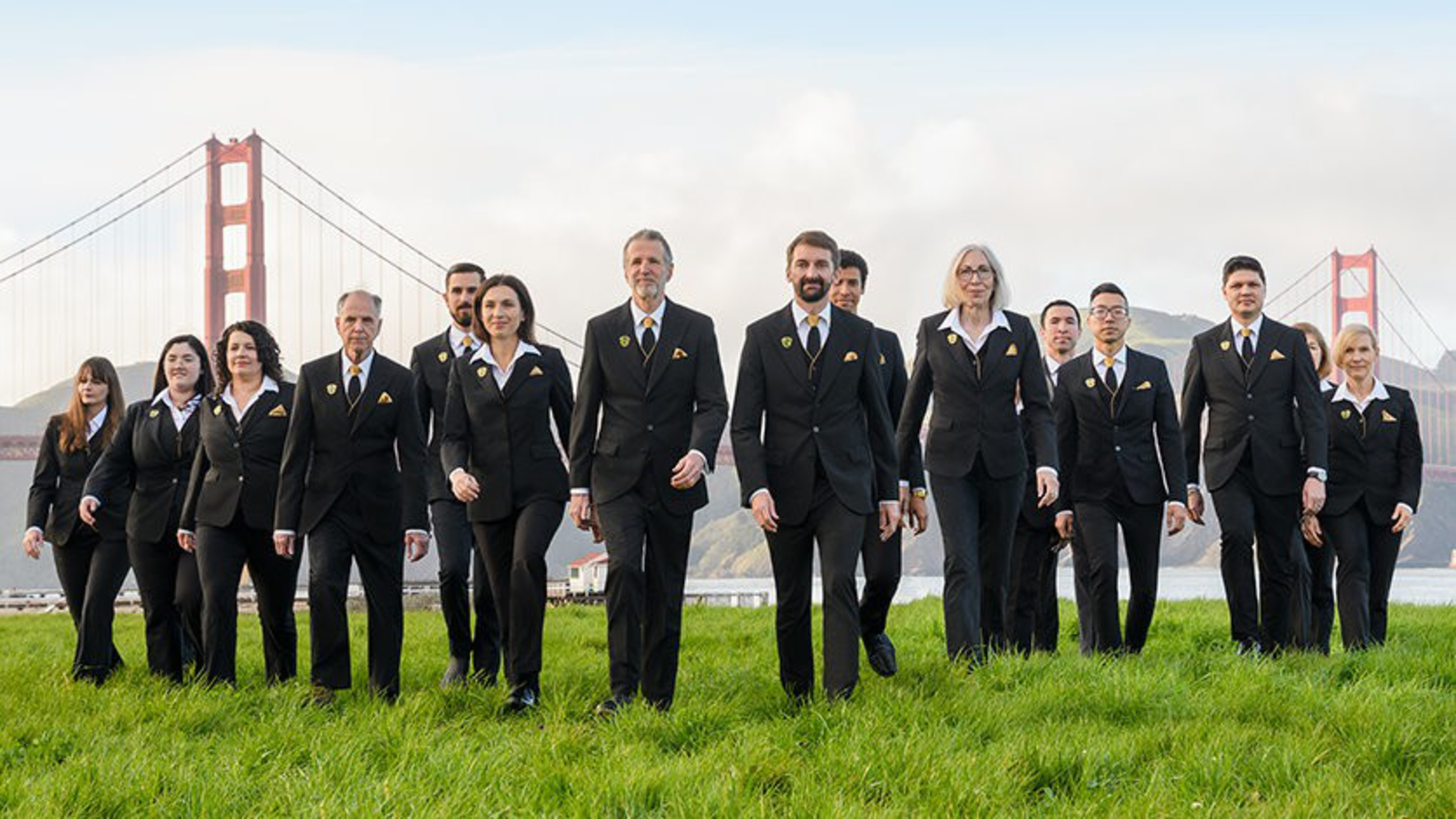A group of people in black suits with yellow accents walk on grass, with the Golden Gate Bridge visible in the background on a cloudy day.