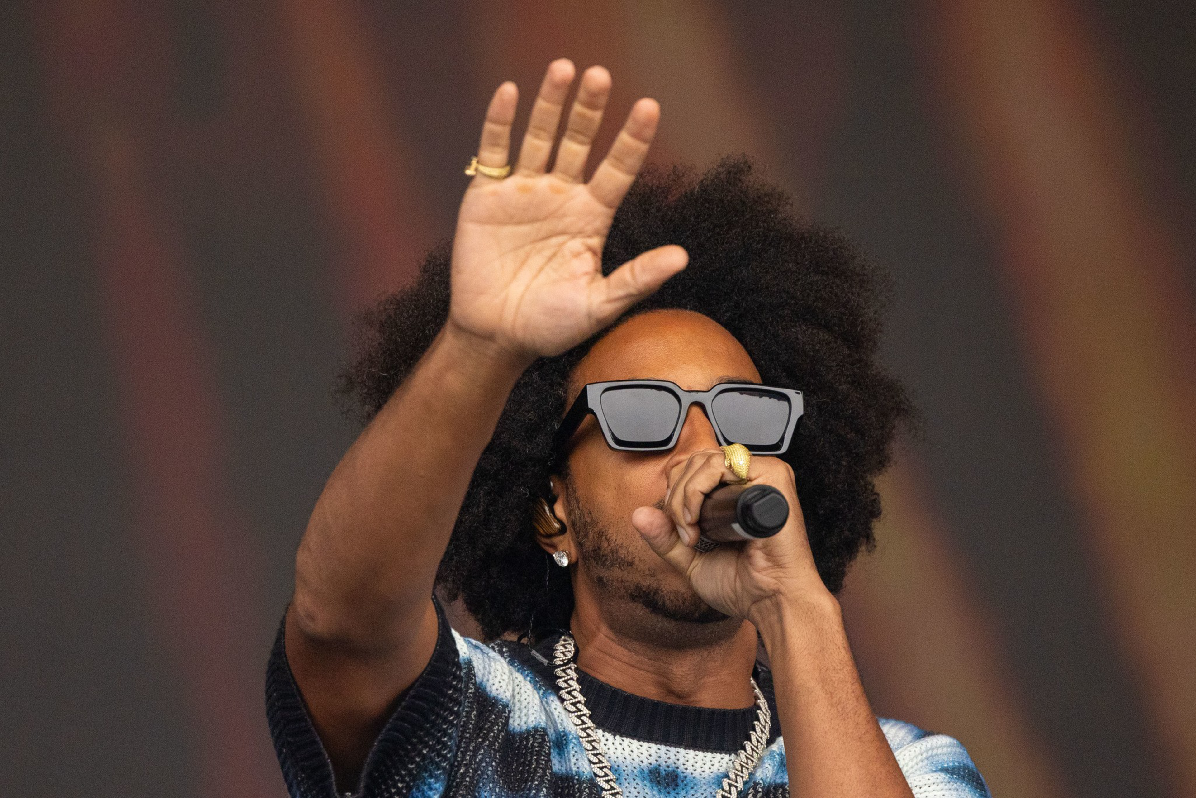 A man with an afro wearing large black sunglasses and a chain necklace holds a microphone near his mouth with one hand while raising his other hand.