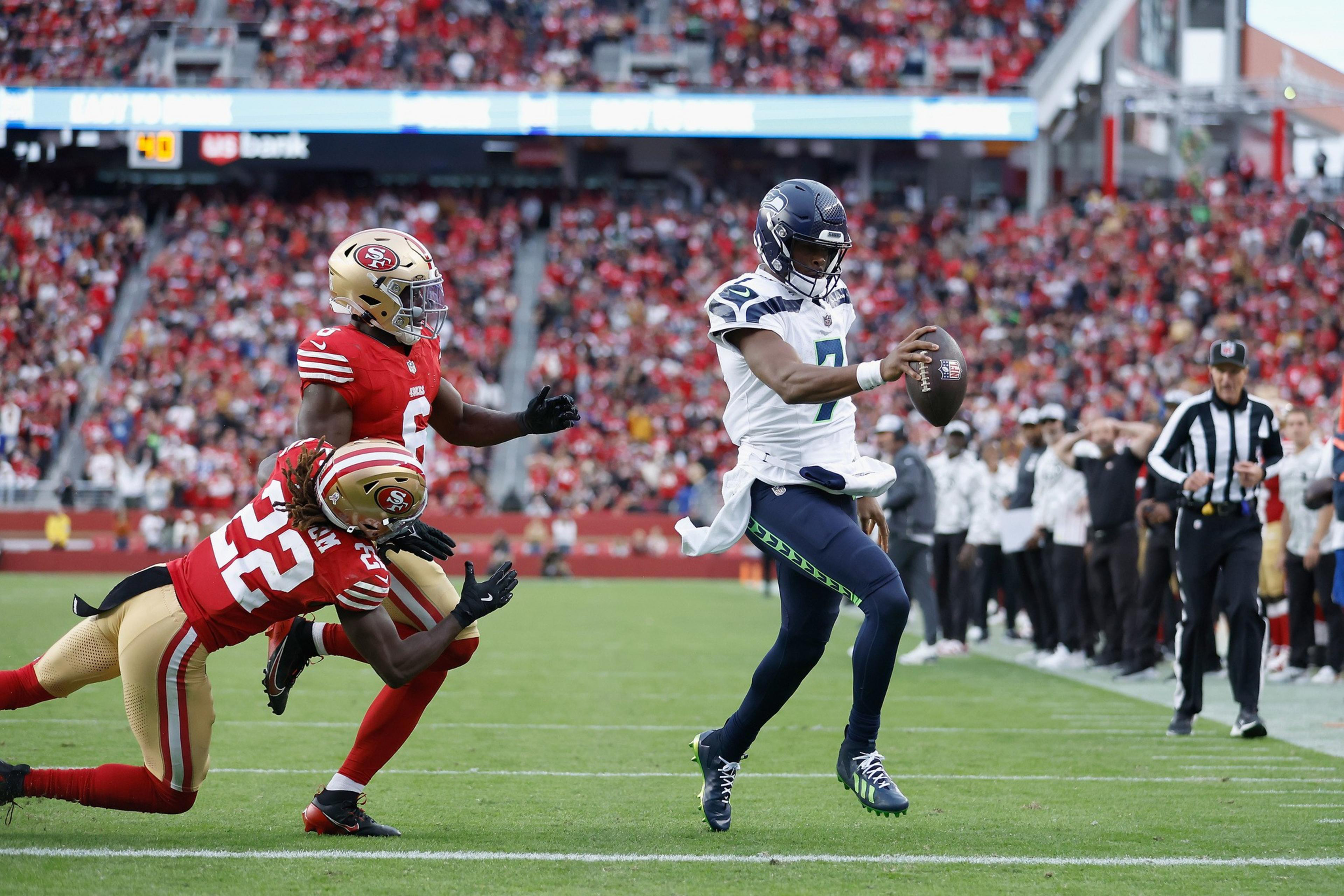 A football player in a white and blue uniform is running with the ball near the sideline, pursued by two players in red and gold uniforms from the opposing team.