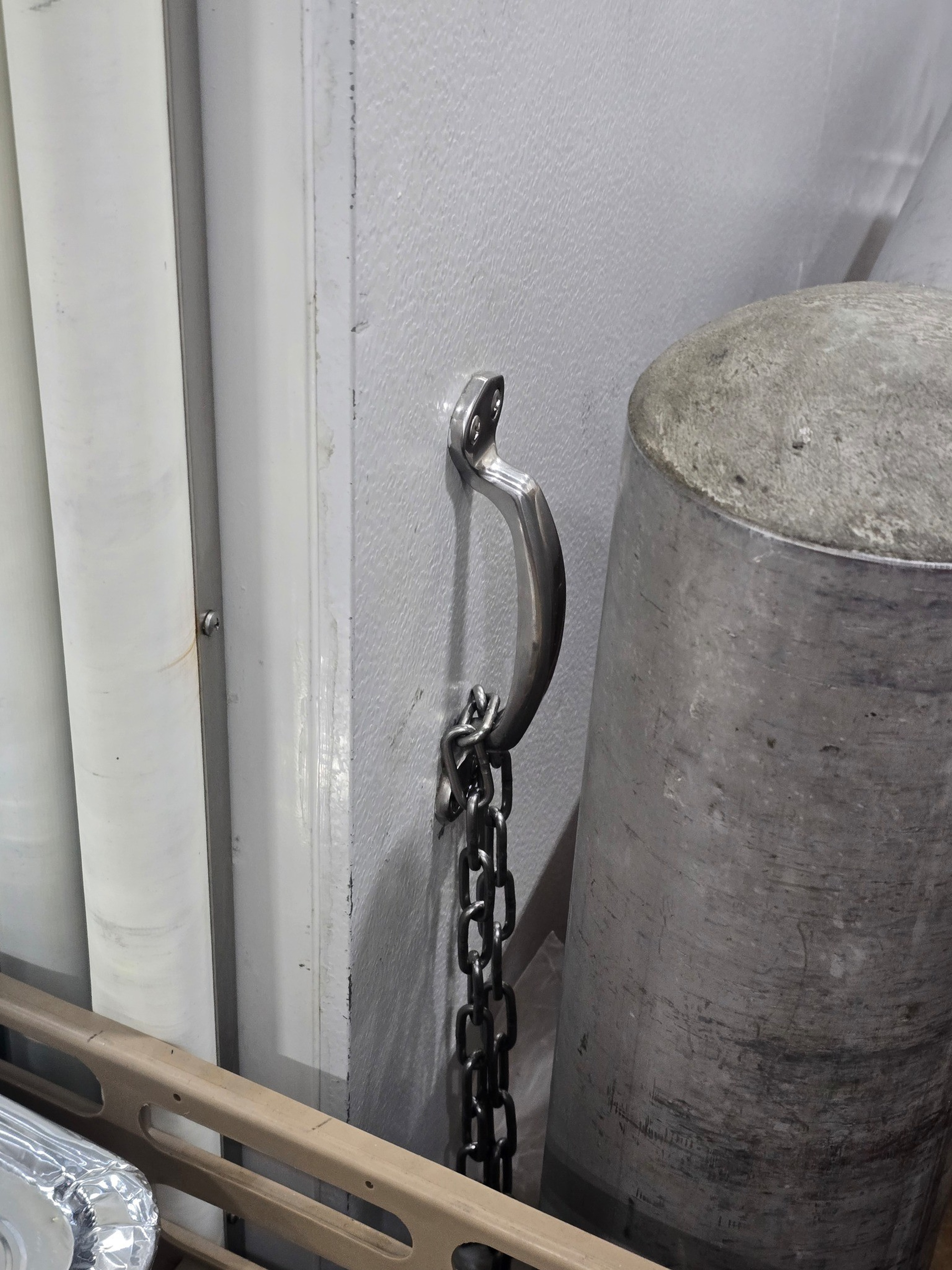 A silver curved handle is mounted on a white wall with a heavy black metal chain hanging from it, next to a large, cylindrical metallic object.