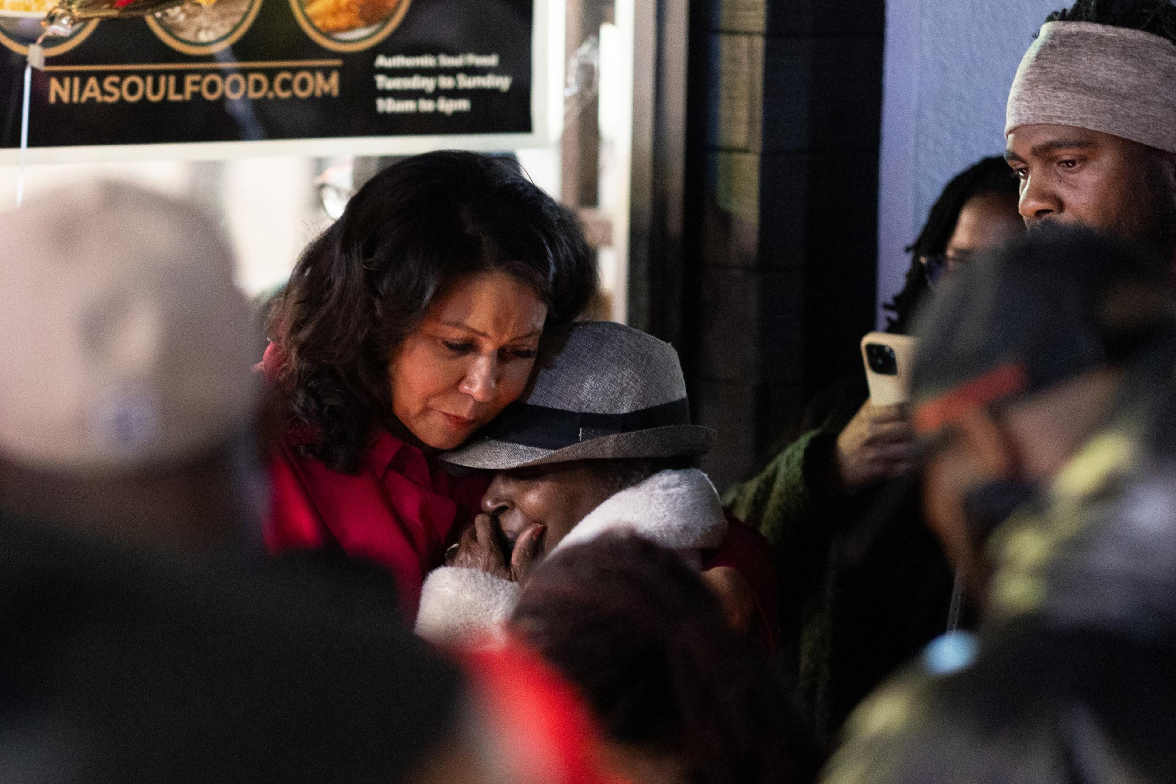 A woman in a red coat comforts another woman wearing a gray hat outside a restaurant. They are surrounded by a group of people, some holding phones.