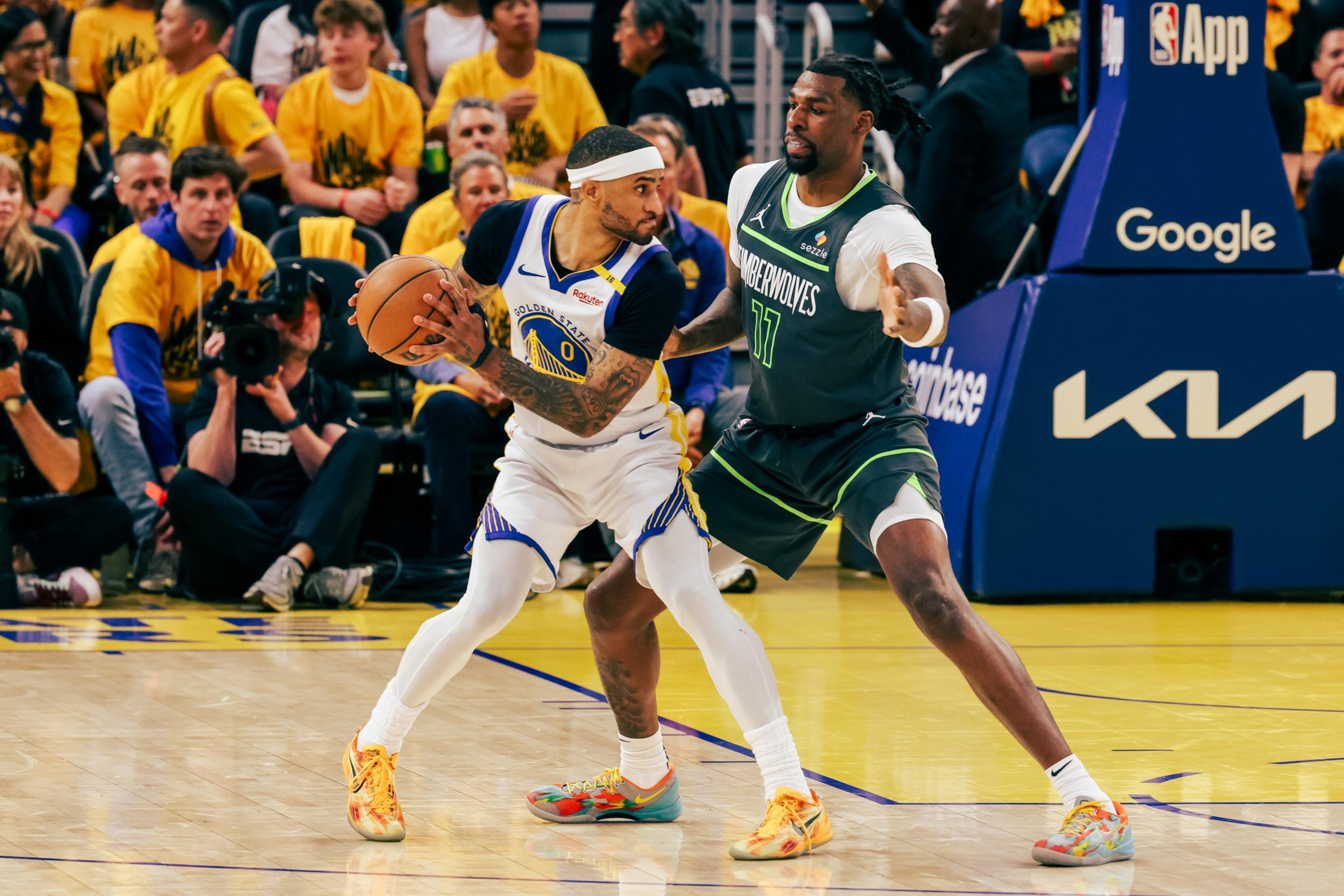 A basketball player in a white and yellow uniform holds the ball, guarded closely by a player in a gray and green uniform. Spectators and a cameraman are visible.