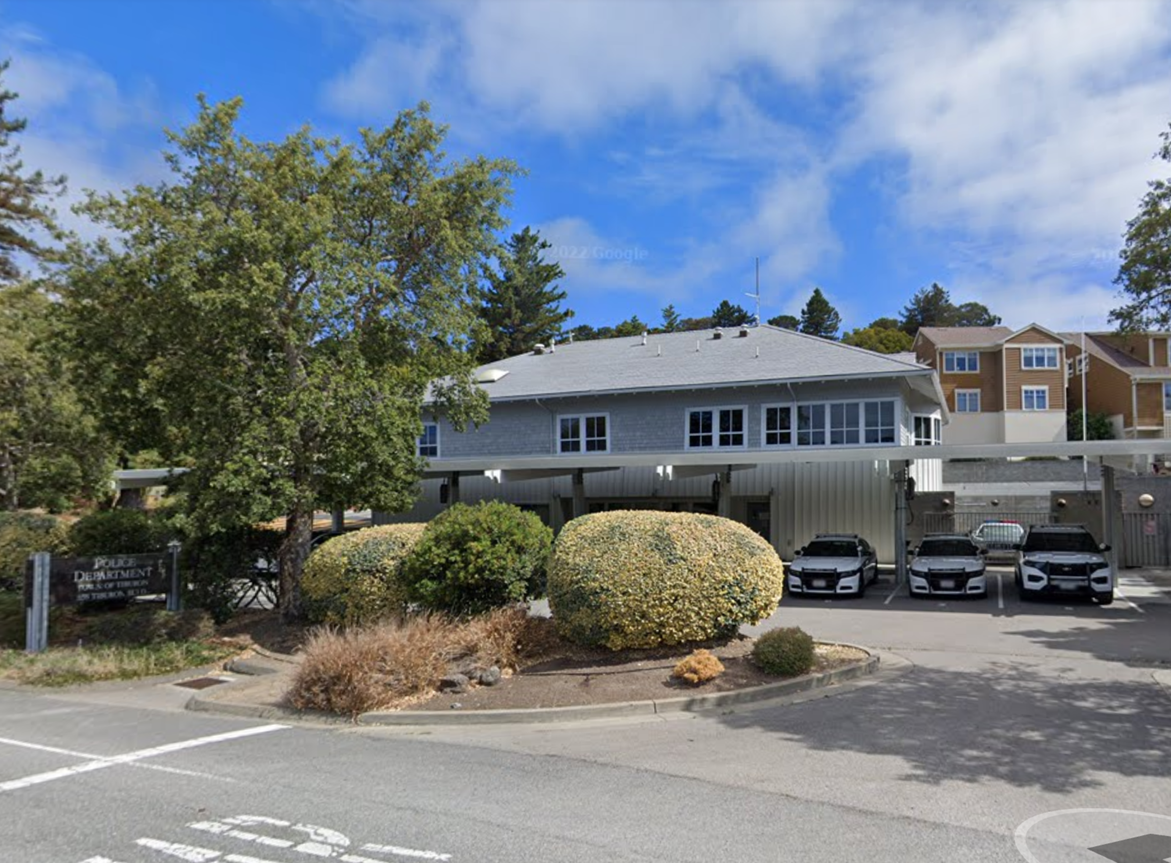 A two-story police department building with a gray roof, surrounded by bushes and trees, and four parked police cars in front under a blue sky.