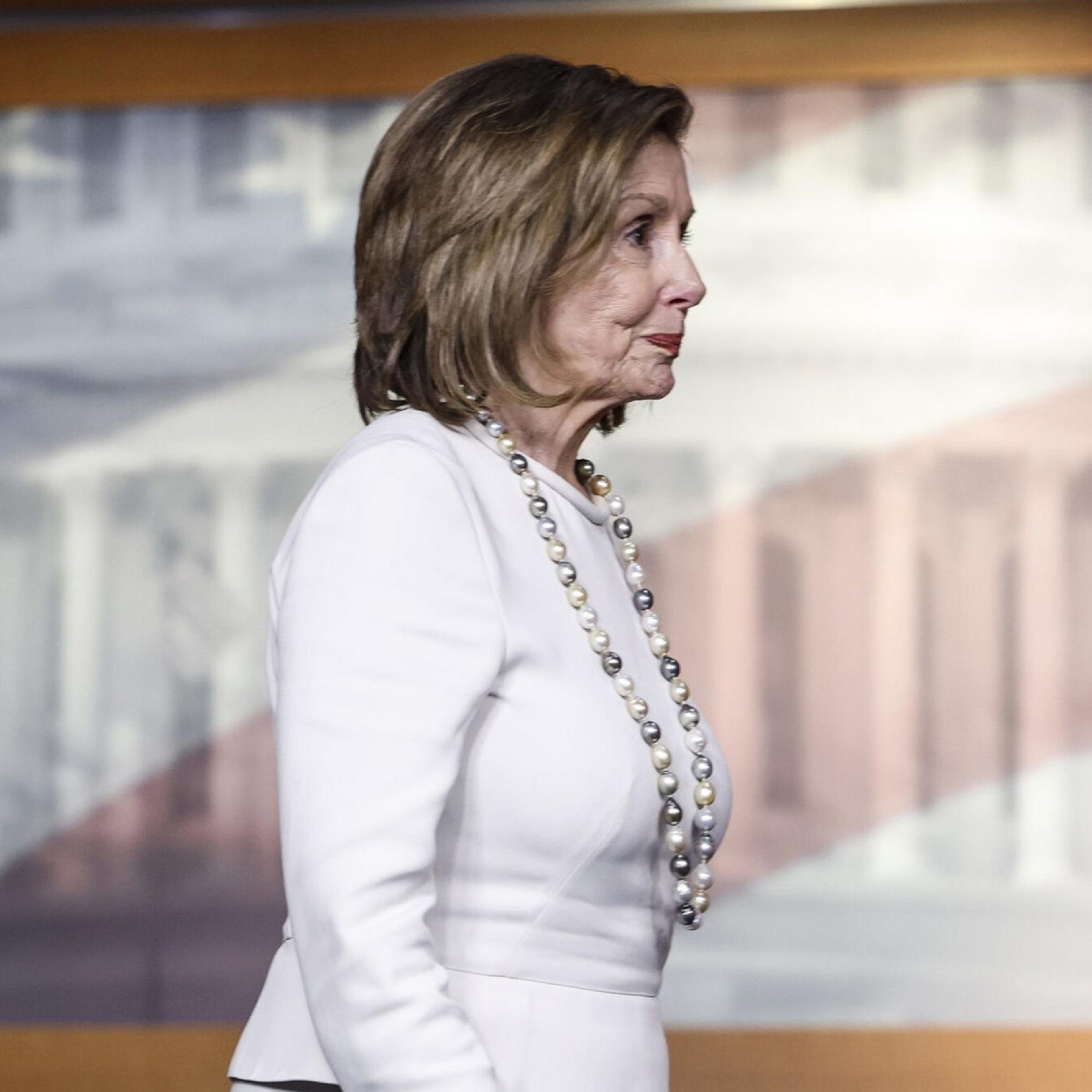 A woman in a white suit and pearl necklace stands sideways in front of a backdrop featuring a blurry image of the U.S. Capitol Building and American flag.