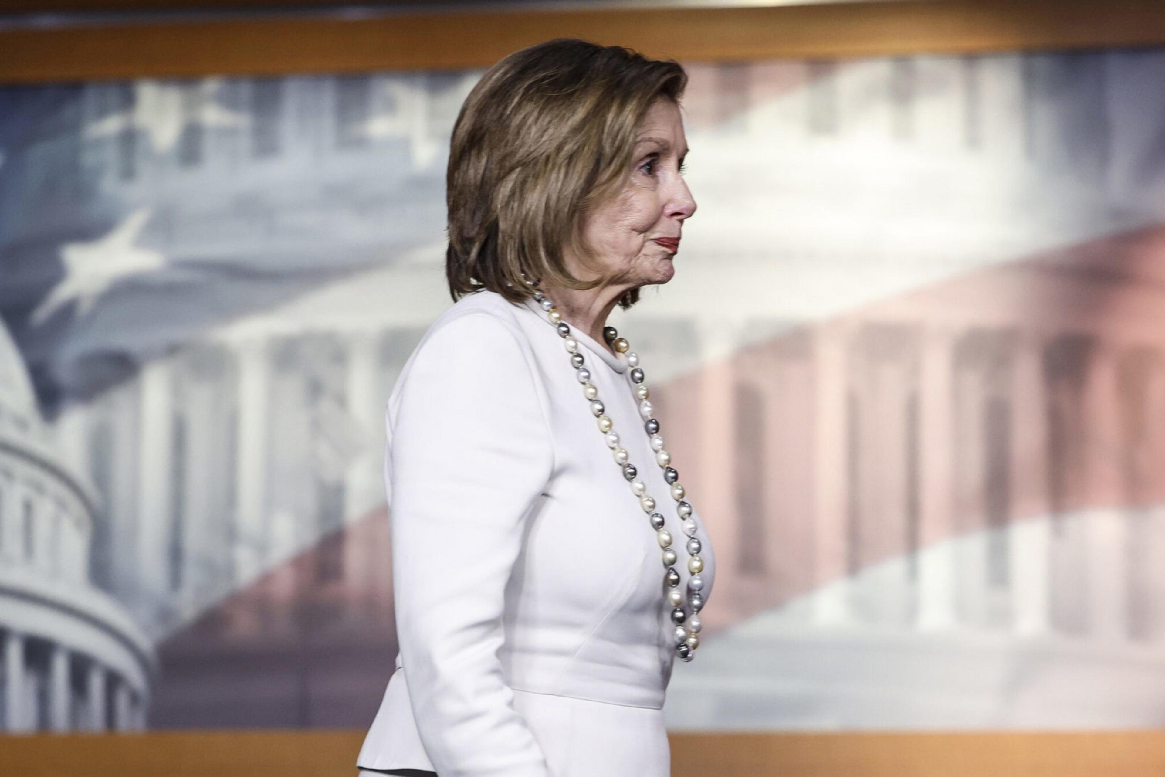 A woman in a white suit and pearl necklace stands sideways in front of a backdrop featuring a blurry image of the U.S. Capitol Building and American flag.