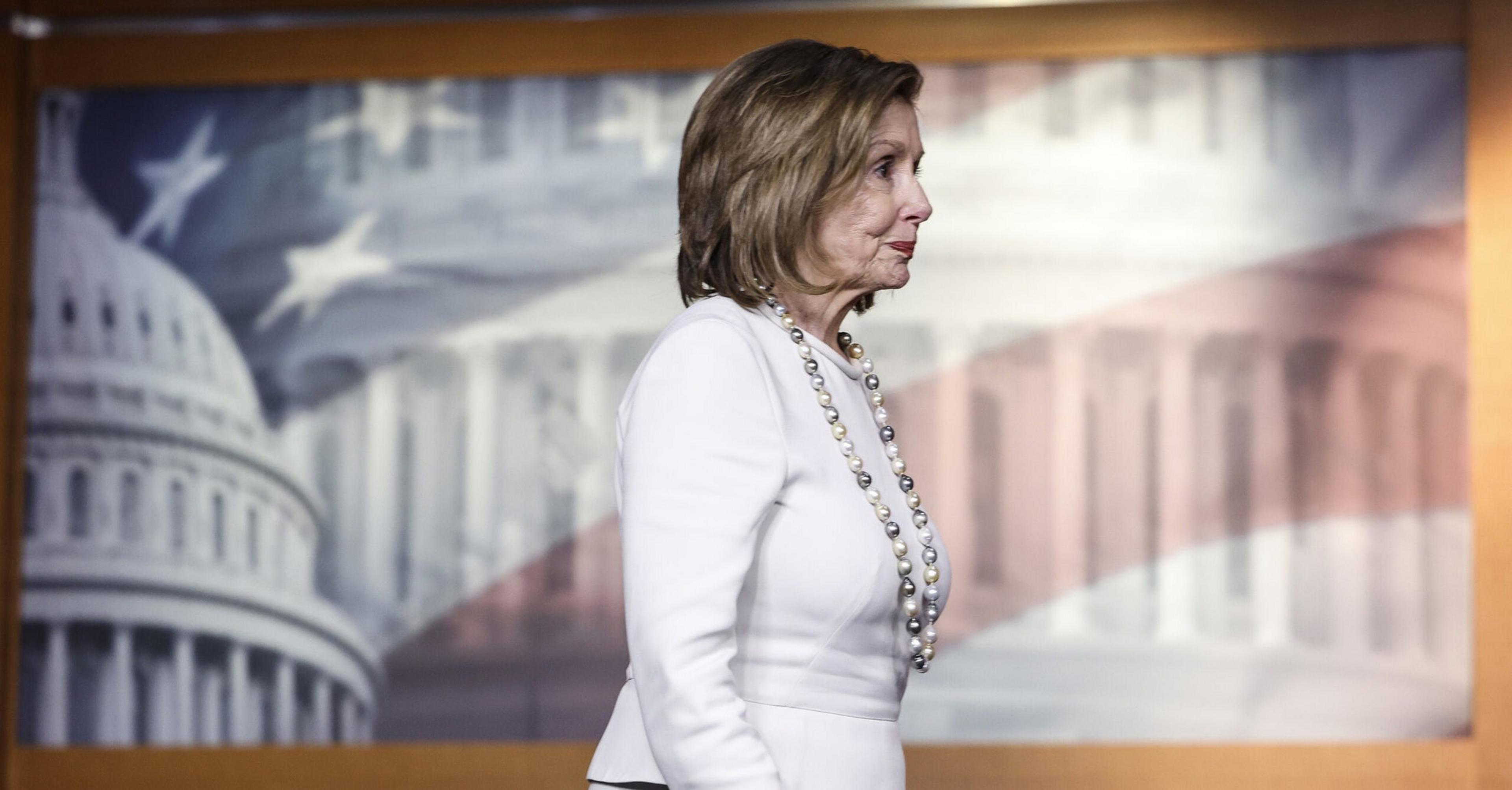 A woman in a white suit and pearl necklace stands sideways in front of a backdrop featuring a blurry image of the U.S. Capitol Building and American flag.