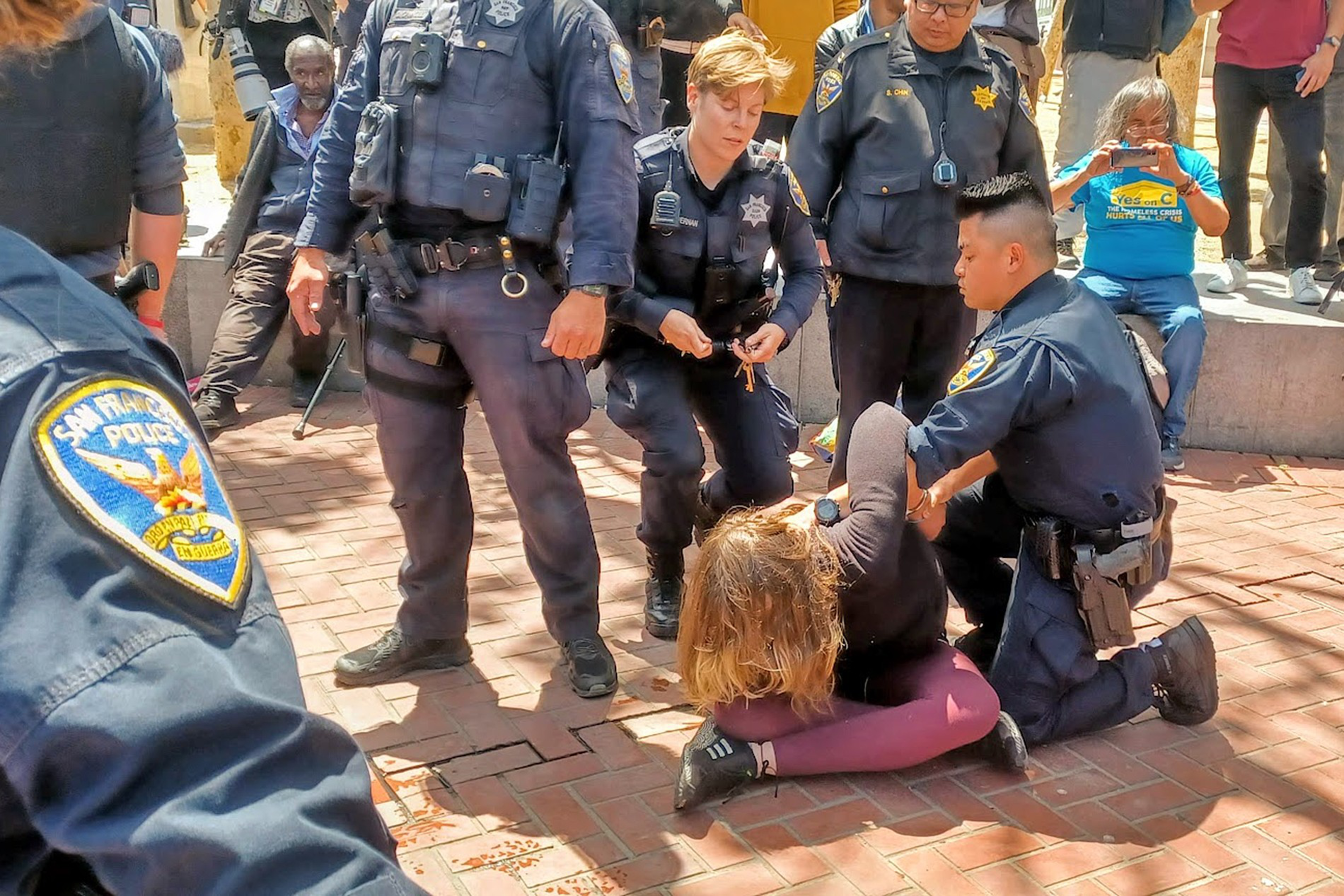 Several police officers restrain and handcuff a person lying face down on a brick sidewalk while bystanders watch nearby.