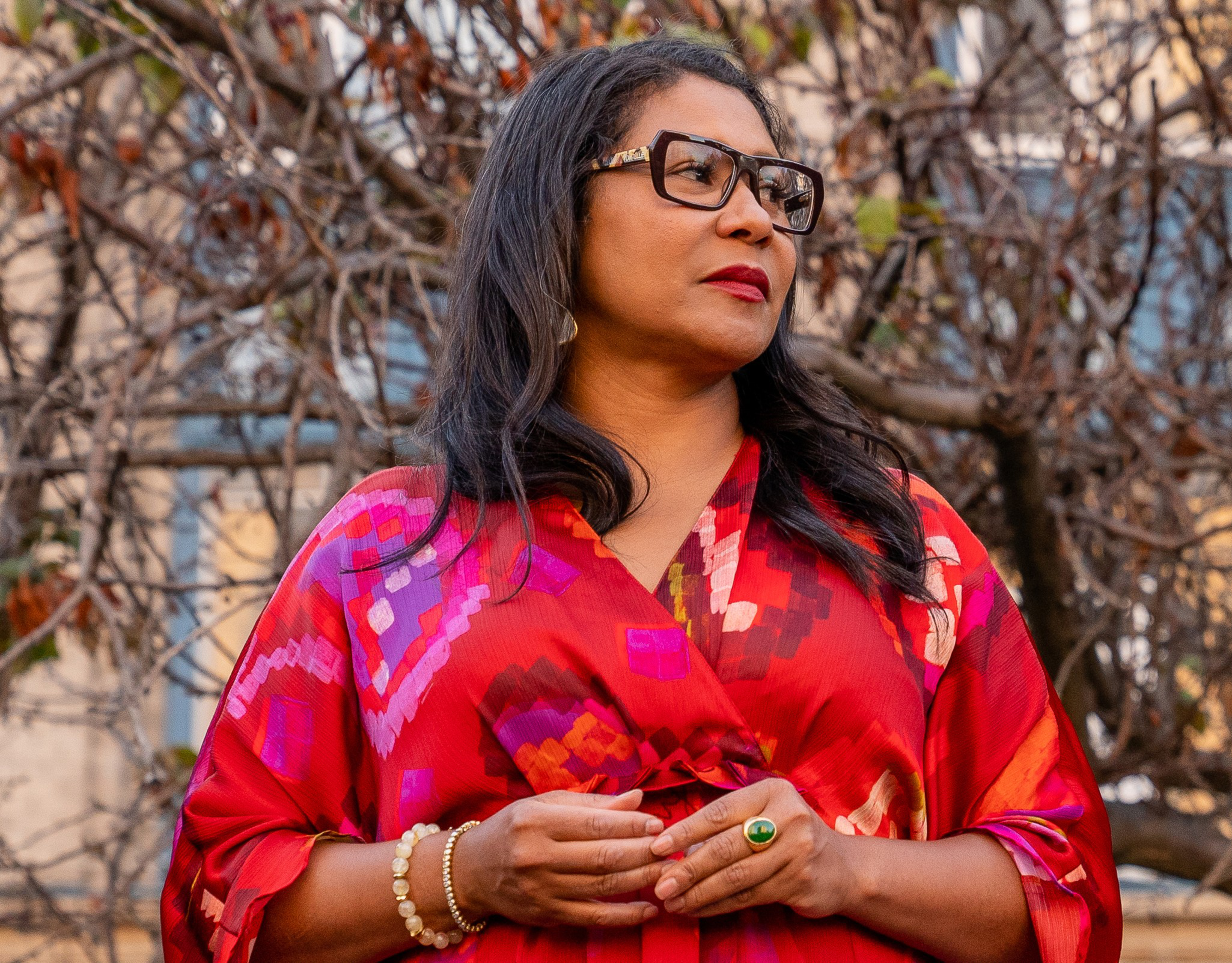 A woman with long dark hair and glasses wears a red patterned dress, a green ring, and bracelets, standing outdoors with leafless branches behind her.